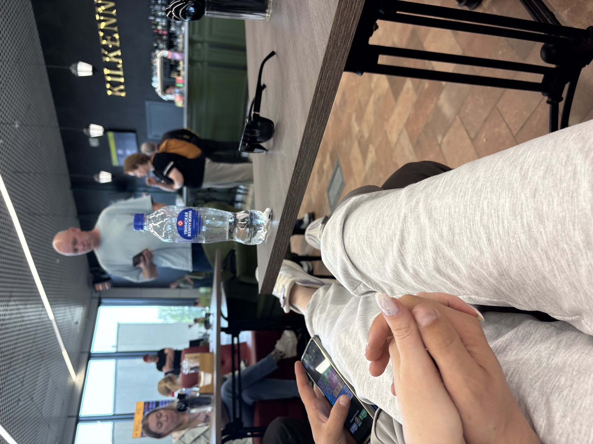 The photograph captures a scene at what appears to be a bar or restaurant, likely at an airport or transportation hub, identified by the sign reading "KILKENNY." A water bottle is prominently placed on the table, alongside a pair of sunglasses. A person, identified as holding a smartphone, is in focus. In the background, there are several individuals in transit, some seated and some standing. The environment appears to be a waiting area or a casual dining space, situated possibly in Новое Мартьяново, Russia, given the inscription on the water bottle.