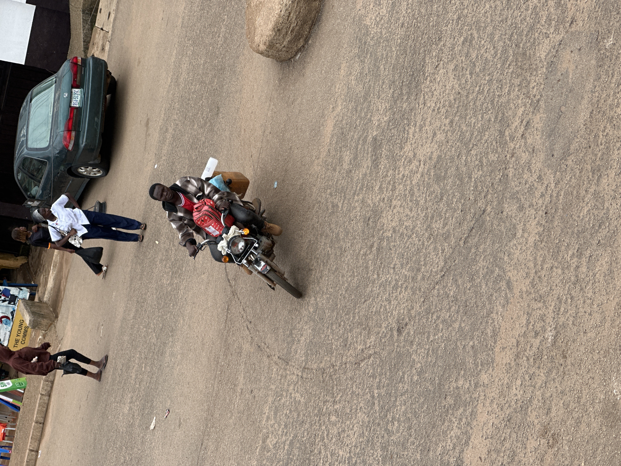 In Bukuru, Nigeria, a man is riding a motorcycle down a dirt road. He is wearing a plaid jacket over a red shirt and dark pants. A red bag is strapped to the back of the motorcycle. To the left of the motorcycle, a person wearing a dark hoodie and black pants is walking away from the camera, holding a black umbrella. Further in the background, a dark green sedan is parked on the right side of the road. Two women are standing near the sedan. One woman, wearing a white shirt and dark pants, is looking towards the motorcycle. The other woman, wearing a black shirt, is standing behind her. The road appears to be unpaved and dusty.
