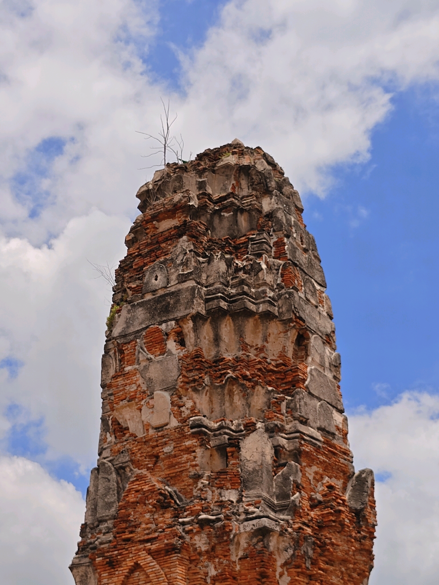 A close-up, low-angle shot captures the weathered, decaying brickwork and stone structure of a Buddhist temple spire against a bright blue sky with scattered white clouds. The spire, located in the Historic City of Ayutthaya, Thailand, shows significant erosion and damage. Reddish-brown bricks are visible in many sections, interspersed with grey stone architectural elements that appear chipped and broken. Some openings and indentations in the structure suggest past decorative features or structural damage. A few thin, bare branches of a plant are growing from the top and side of the spire, indicating nature's reclaiming of the ancient ruin. The lighting suggests it is daytime, with clear visibility.