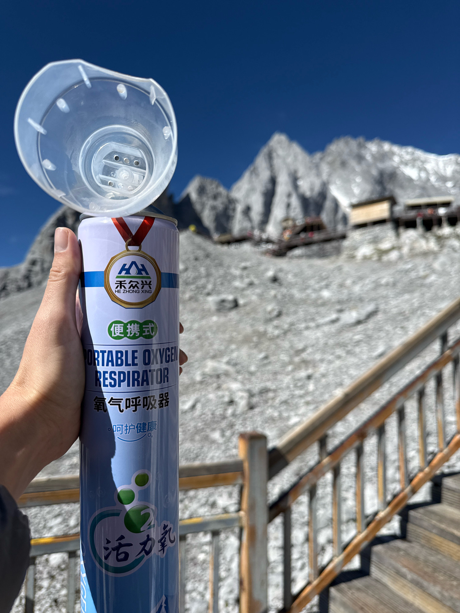 A person's hand is holding a light blue aerosol can with a clear plastic mask attachment. The can has a label with text in English and Chinese, including "PORTABLE OXYGEN RESPIRATOR" and "活⼒氧". The background is a blurry mountainous landscape under a clear blue sky, with wooden stairs and railings visible in the foreground. The scene appears to be in a high-altitude location, possibly in Yulong Nakhi Autonomous County, China, given the presence of a portable oxygen respirator.