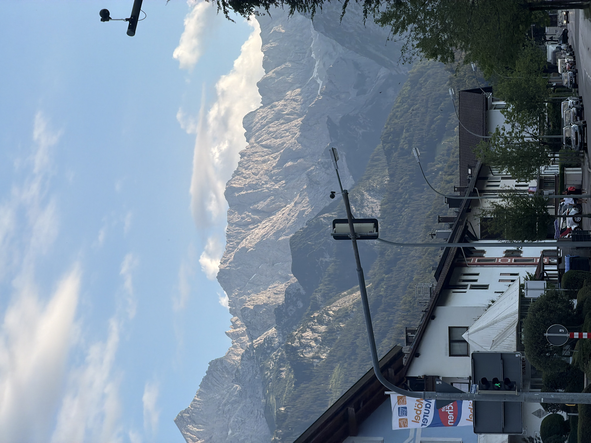 This is a vertical, eye-level, outdoor, medium-shot photograph taken on a street in Garmisch-Partenkirchen, Germany. In the background, a large mountain range with rocky, jagged peaks and tree-covered slopes dominates the upper portion of the frame. The sky above is a pale blue with scattered white clouds. In the middle ground, a street lined with several buildings is visible. These buildings appear to be commercial or residential, with white and light blue facades, dark roofs, and windows. A traffic light with a green pedestrian signal is prominent in the left foreground. Several cars are parked along the street. In the foreground, trees and a street lamp are also visible. The overall scene depicts a town nestled at the foot of a mountain range.