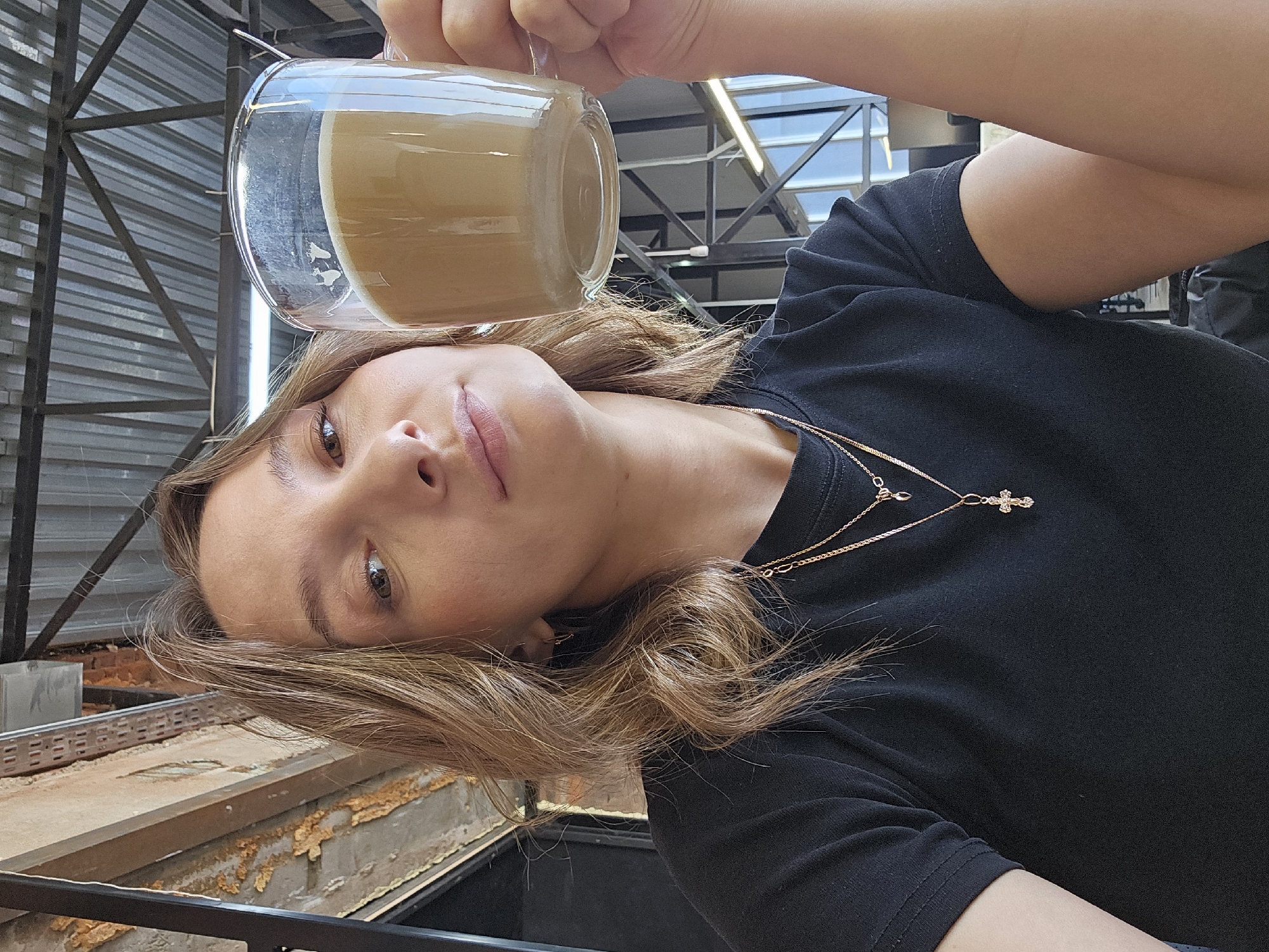 A woman in a black shirt holds a glass mug filled with a light brown liquid. She wears a layered gold necklace, the lower chain adorned with a cross pendant. Her brown hair is styled in loose waves. The background includes a metal structure, a gray concrete support, and what appears to be a section of distressed wooden paneling. The image orientation is rotated. The location is Chaikovsky, Russia.
