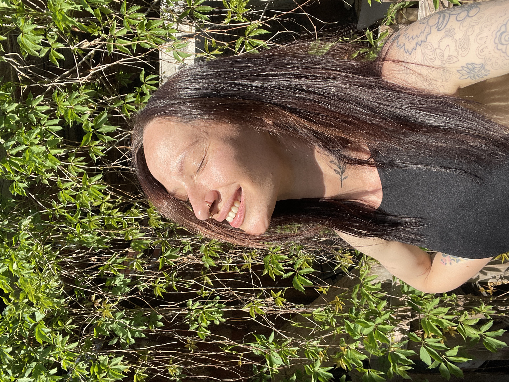 A woman is lying on the ground in an area with green leafy vegetation, possibly a garden or a park. She is wearing a black top and has tattoos on her neck and arm. The woman has dark brown hair and is smiling with her eyes closed, facing the sun. Her head is slightly tilted to the left. The background consists of dense green plants with a wooden fence visible behind them. There is no specific information to ascertain the location as Oktyabr'skii, Russia, but based on the provided visual information, the scene appears to be an outdoor setting in a sunny environment.