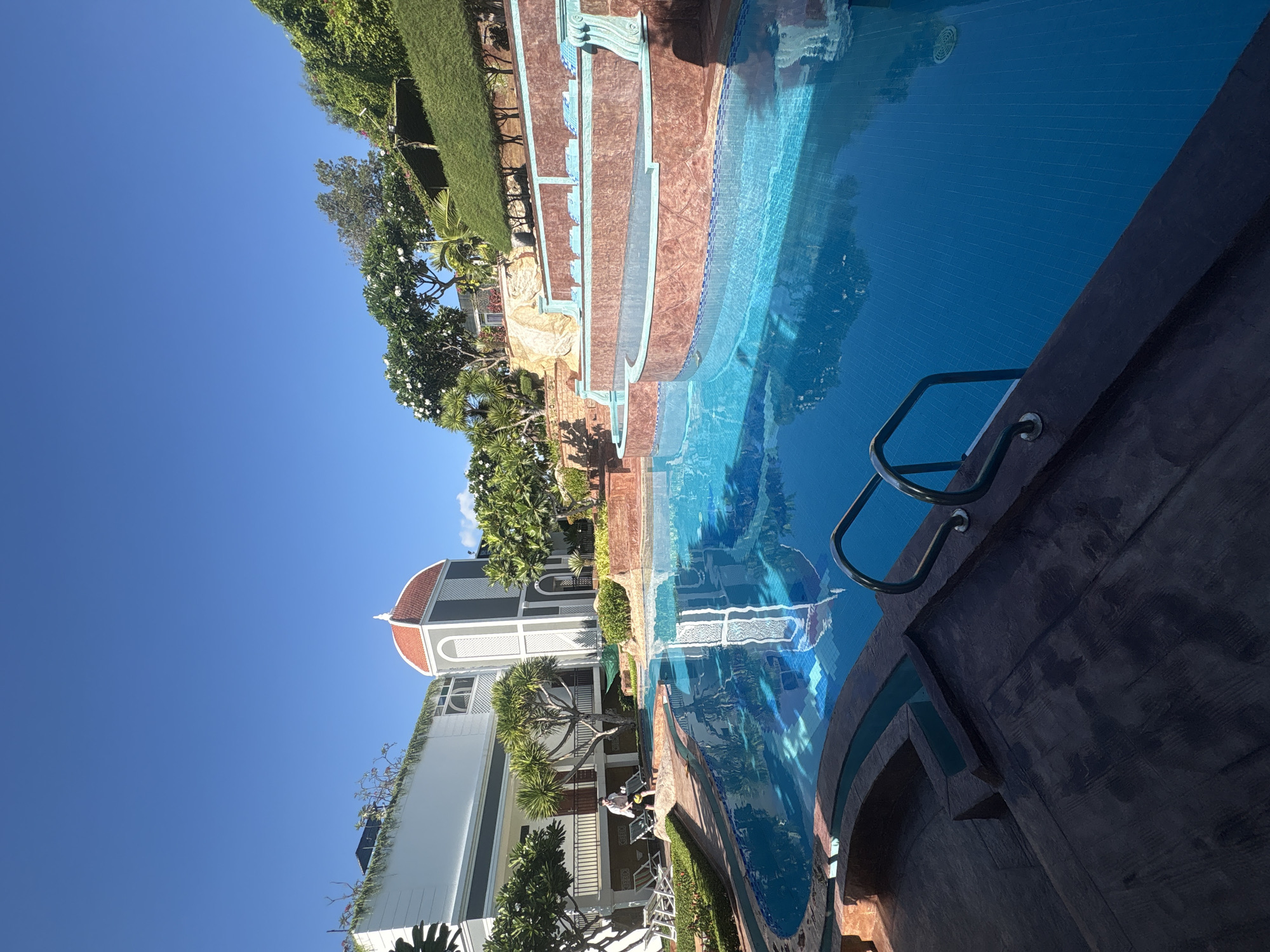 The image captures a swimming pool area at a resort in the Bac Nha Trang Ward of Nha Trang, Vietnam, under a clear blue sky. A multi-level pool features blue-tiled water. A dark-colored edge with a metal ladder defines the pool's edge. Adjacent to the pool, there is landscaped greenery, including trees and grass. A building with a red-domed structure is seen in the background. Two figures appear to be sitting on chairs on a balcony of the building. The architecture incorporates a blend of modern and classical elements.