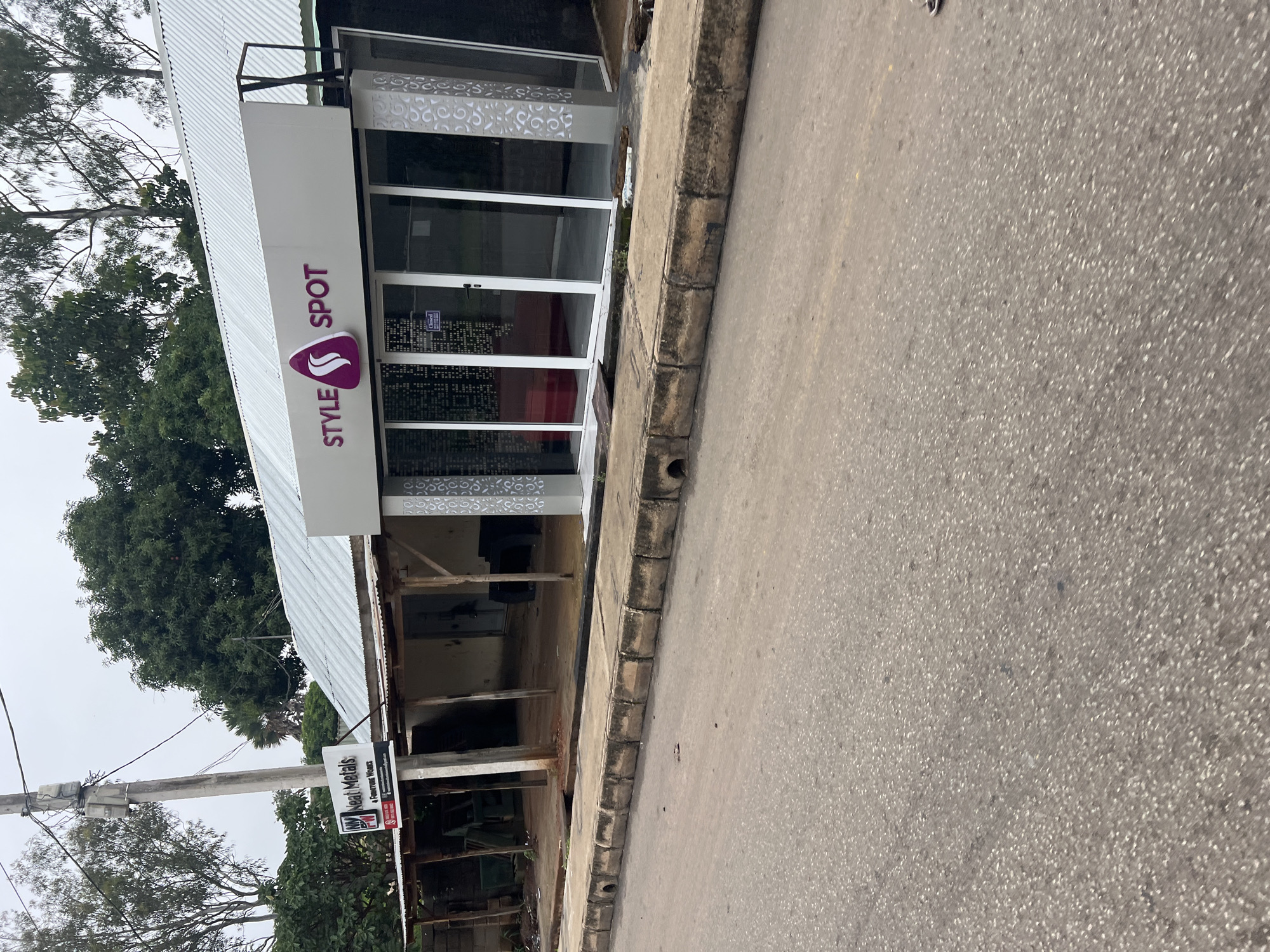 This is a low-angle, vertically oriented exterior shot of a commercial building in Jos, Nigeria. The building appears to be a shop, with a prominent sign that reads "STYLE SPOT" in purple lettering, accompanied by a stylized purple logo. The storefront features large glass windows and doors.
To the left of the "STYLE SPOT" sign, there's a glimpse of the building's corrugated metal roof and some surrounding greenery, including large trees. Below the main shop area, the building seems to be under some form of construction or renovation, with exposed wooden beams and unfinished walls visible.
A separate, smaller sign hangs on a pole in front of the building, which reads "Neat Metals" and "A Corrent Works" with a phone number and a red "WhatsApp" icon. This suggests the presence of another business, possibly related to metalwork or construction services.
The pavement in front of the building is cracked and weathered, with a concrete curb bordering the road. The road itself is paved and appears to be a typical street, with some debris visible. The overall atmosphere suggests a slightly overcast day, possibly in the morning or late afternoon, given the muted lighting. There are no people visible in the frame. The scene depicts a commercial area with a mix of active and under-construction businesses.