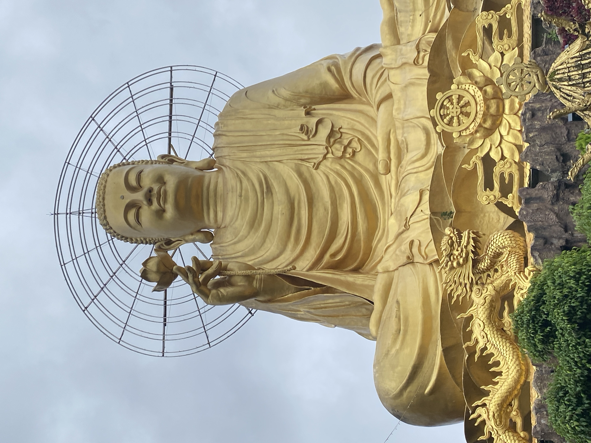 This is a medium-wide shot of a colossal golden Buddha statue, seated in a meditative pose. The Buddha's right hand is raised, holding a delicate lotus flower, while his left hand rests in his lap. A circular, metallic halo or mandorla surrounds the Buddha's head. The statue is adorned with intricate golden details, including a dragon sculpture to the left of the base, and a Dharma wheel motif to the right, nestled within floral and cloud-like designs. The base of the statue appears to be constructed from rocky material, with lush green foliage visible in the foreground. The sky is overcast with a pale gray hue. This statue is located at the Truc Lam Zen Monastery in Lâm Viên - Đà Lạt, Vietnam.
