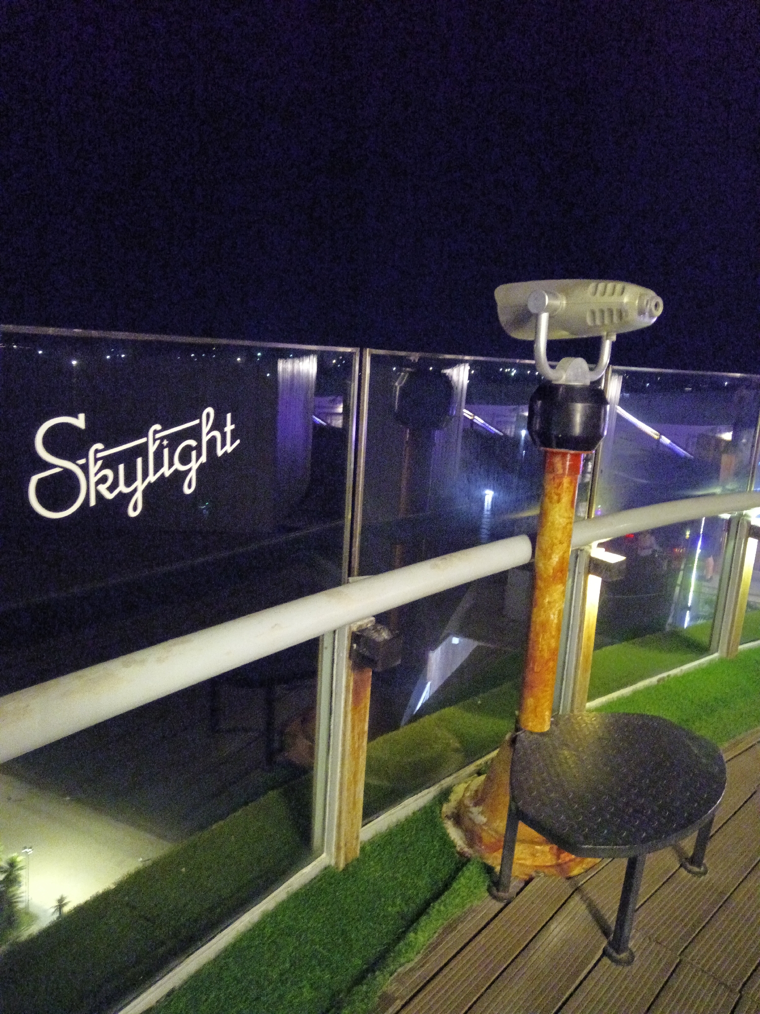 At night, the Skylight rooftop bar in Nha Trang Ward, Vietnam, features a coin-operated telescope mounted on a circular metal platform. The telescope is situated on a raised deck covered in artificial grass, behind a glass barrier. The bar's name, "Skylight," is displayed in white cursive lettering on the glass. Below, the dark cityscape is partially visible. The sky above is dark.