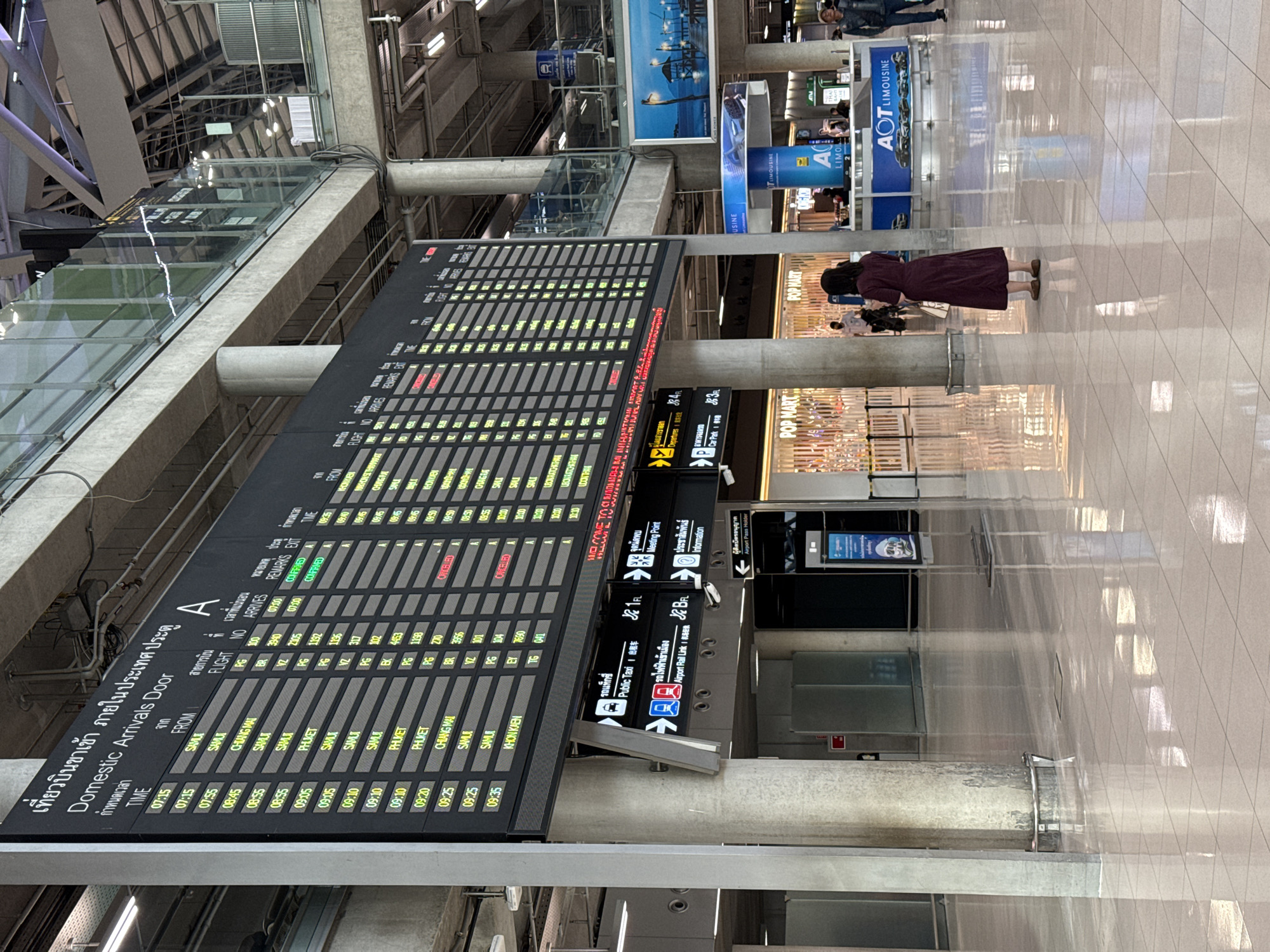 An interior shot captures a section of a busy airport terminal. A large digital departures and arrivals board dominates the upper portion of the frame, displaying flight information in both Thai and English. The board is mounted on a large concrete pillar. Below the main board, directional signage indicates "Public Taxi," "Airport Rail Link," "Meeting Point," and "Information." To the right, a person, presumably a woman, stands facing away from the camera, wearing a dark maroon dress. In the background, various kiosks and shops are visible, including "POP MART" and an "AOT LIMOUSINE" counter. The floor is polished, reflecting the overhead lights and architectural elements. The environment appears to be a clean, modern airport concourse. The location is identified as Nong Prue Subdistrict, Thailand.