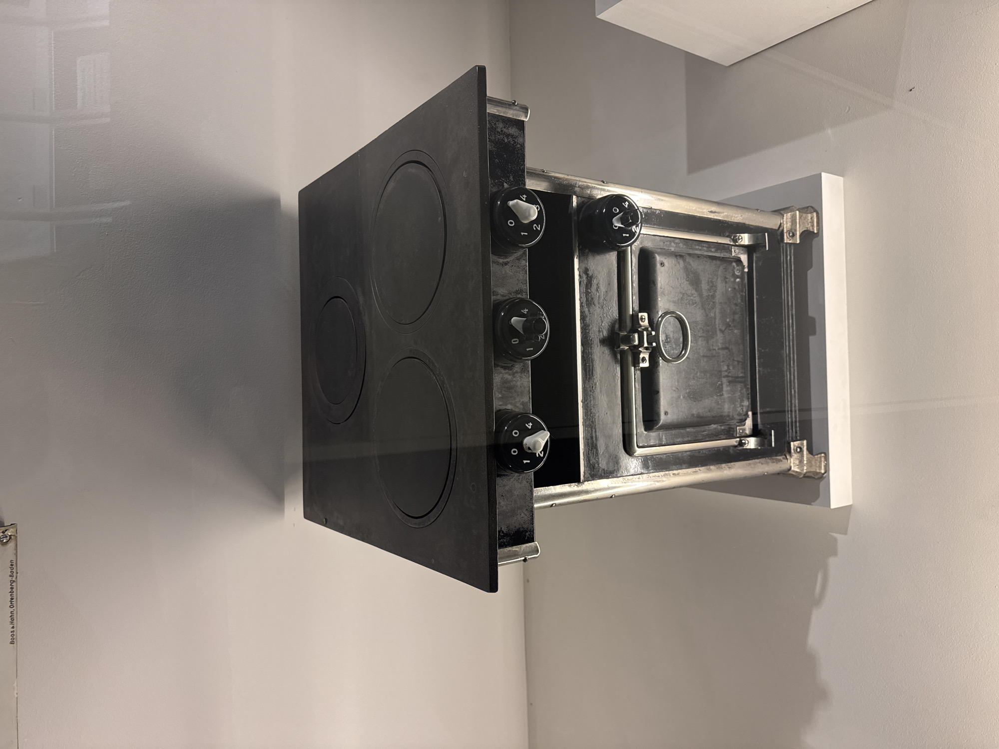 This media file shows a close-up, tilted shot of a vintage black stove with a metal frame, positioned against a plain, light-colored wall. The stove has a flat top with three circular heating elements, and a small oven door is visible on the right side. Three circular control knobs with white indicators are mounted on the front panel of the stove, below the heating elements. These knobs appear to be set to different levels, indicated by numbers ranging from 0 to 4.
The lighting in the image is diffused, casting soft shadows on the wall, suggesting an indoor setting, possibly a museum or an exhibition space, given the clean presentation. The overall tone is neutral, focusing on the object itself. The environment appears to be dimly lit, which highlights the texture of the wall and the details of the stove.
There is text visible on a vertical metal strip on the far left of the image, which reads "Boos + Hohn, Ortenberg, Baden". This text provides a manufacturer or origin cue. The image orientation is slightly tilted, giving a dynamic perspective of the stove. The scene does not depict any people or specific activities, but rather presents a static display of a historical kitchen appliance. The location context is Bern, Switzerland, but this information is not visually evident from the image itself.