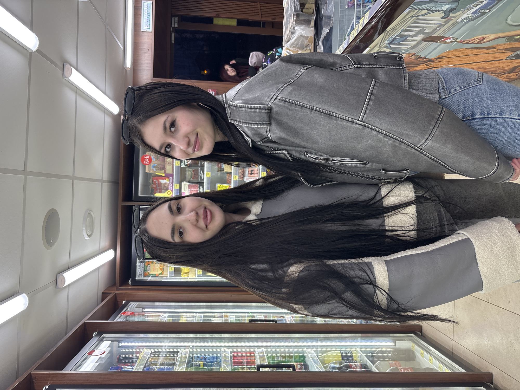 Two young women are captured in a casual moment, posing for a photo inside what appears to be a Russian supermarket or convenience store in Фоки. Both women have long, dark hair and are smiling warmly at the camera.
The woman in the foreground is dressed in a faded gray denim-style jacket and light blue jeans, with black sunglasses resting on her head. Slightly behind her, the second woman wears a distinctive gray jacket with wide white and gray stripes, featuring a fuzzy white collar and cuffs.
The setting is characterized by numerous refrigerated display cases lining the walls, filled with a variety of products, likely food items and beverages. Above these cases, the walls are light-colored, and the ceiling is fitted with long fluorescent tube lights and circular recessed lighting or speakers, illuminating the interior brightly.
Visible text on the product displays includes "K&Б," "Original," and the Cyrillic word "ПРОДУКТЫ" (Products). In the far background, a faint Cyrillic sign, possibly "ВЫХОД" (Exit), can be seen. A reflection in one of the display cases reveals another person and a painted mural depicting a street scene with buildings, a car, and hands. The overall mood of the image is cheerful and relaxed, with both subjects exhibiting positive emotions.