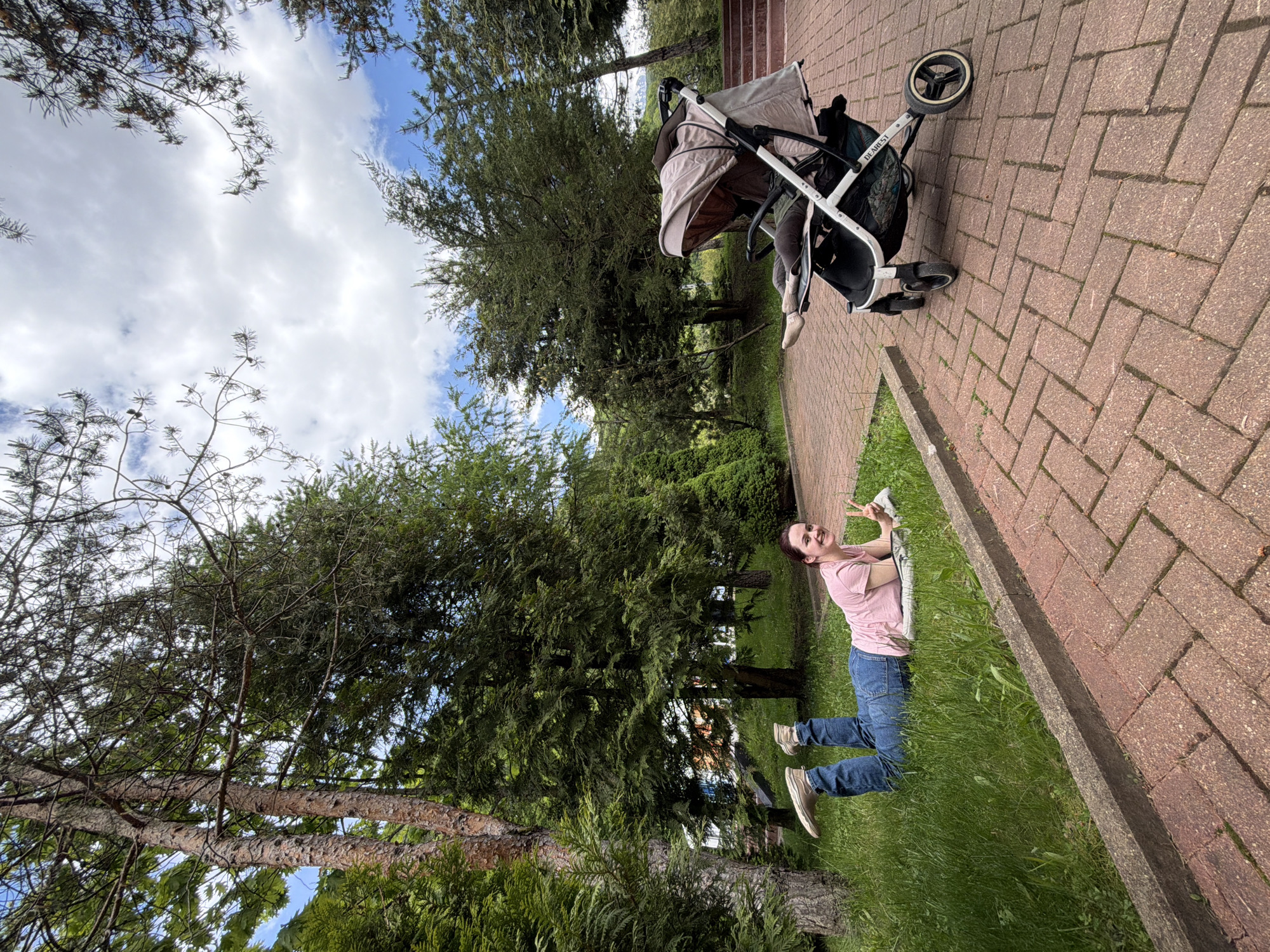 A woman in a pink shirt and blue jeans lies on the grass next to a brick walkway in Estosadok, Russia. She is making a peace sign with her hand. A baby stroller is parked on the walkway nearby. The sky is visible through the trees.