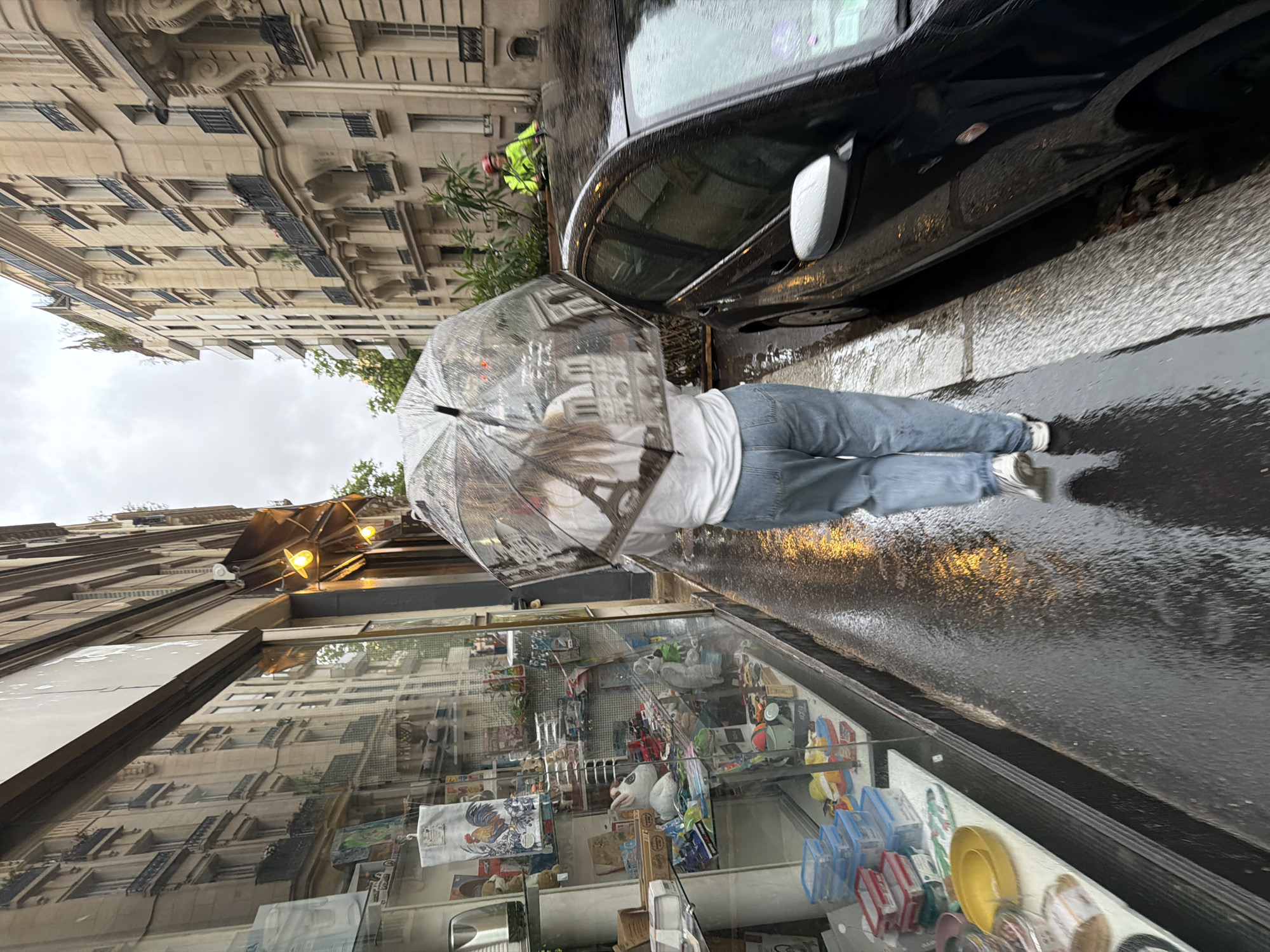 This media file captures a street scene in Paris, France, on a rainy or recently rained-on day. The ground, especially the asphalt, is visibly wet and reflective, indicating recent precipitation. The sky is overcast.
In the immediate foreground, a person walks towards the left, holding a clear, transparent umbrella. They are wearing blue jeans and a white t-shirt, parts of which are visible through the umbrella. The umbrella itself reflects parts of the surrounding buildings and the person's clothing.
To the right of the walking person, a black car is parked along the wet curb. Further down the street, on the far side of the parked car, a second person is visible wearing a bright yellow or lime green jacket and a helmet, possibly on a bicycle or scooter.
On the left side of the frame, a shop window displays various items, including a yellow plate, plastic containers, a white animal figurine (possibly a rabbit or dog), and a decorative cloth with a rooster motif. The shop window also reflects the ornate Parisian buildings across the street.
The background features classic, multi-story Parisian buildings with detailed architecture, including balconies, intricate moldings, and numerous windows with dark shutters. Some vegetation is visible between the buildings and the street. The overall impression is that of a bustling city street enduring a wet day. No clearly legible text is visible in the scene.
