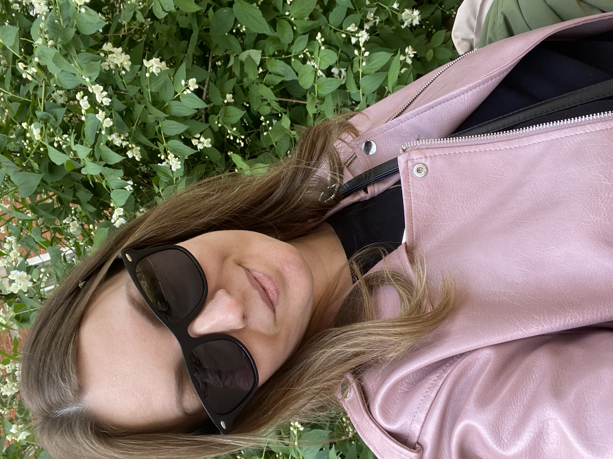 A medium shot depicts a woman in Noviy, Russia, situated amidst vibrant green foliage dotted with small white flowers. She is angled to the left, sporting dark sunglasses. Her light brown hair with blonde highlights falls around her face and shoulder. She wears a pink leather jacket over a black top. The background includes more foliage and a glimpse of what seems to be another person in green.