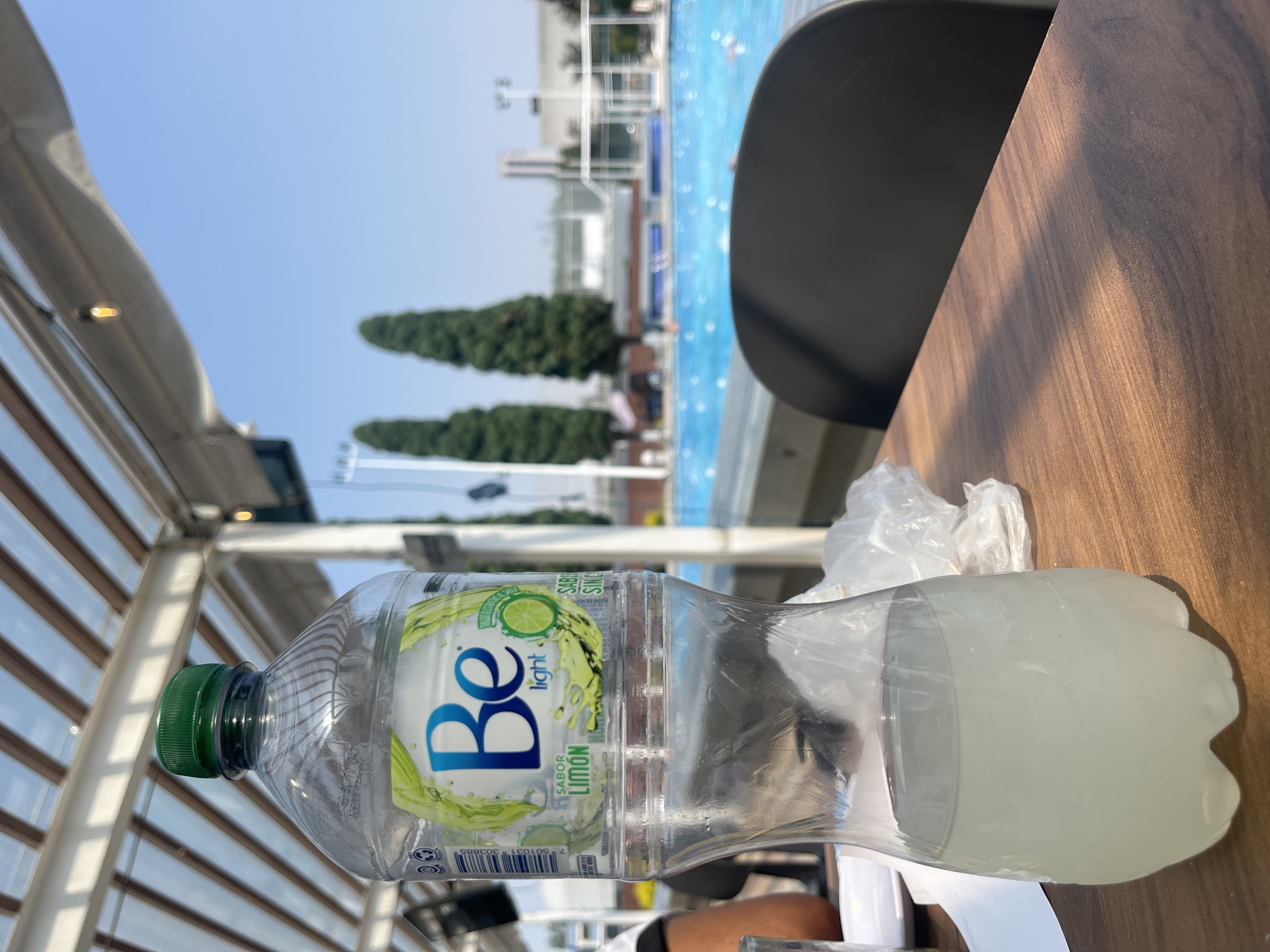 Here is a detailed description of the image:
The image is taken from a slightly elevated perspective, focusing on a scene that appears to be outdoors, possibly near a recreational area. In the foreground, a partially consumed bottle of "Be light LIMON" drink is prominently featured. The bottle has a green cap and is filled with a cloudy liquid. It lies on a wooden table, partially covered by what seems to be crumpled plastic wrap or a plastic bag.
Behind the bottle and table, there is a clear view of a swimming pool. Some people can be seen enjoying the water. The pool is bordered by greenery including tall, cone-shaped trees, and various structures are in the background, including what looks like a building complex.
To the left of the frame, there is an architectural structure with a series of parallel bars, resembling a modern pergola or outdoor canopy. The sky is visible between the structures, indicating it is daytime.