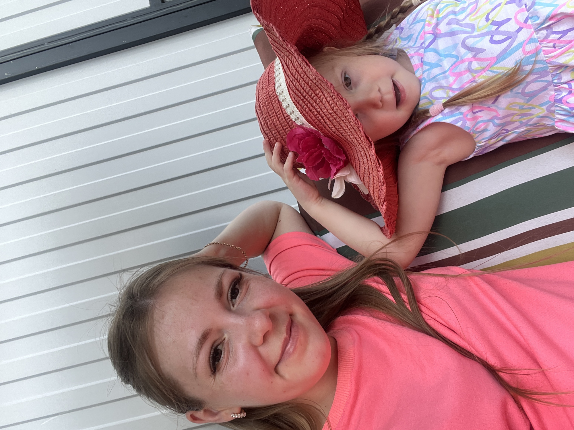 Two females, an adult and a child, are seated on a striped outdoor chair. The adult, on the left, has blonde hair and is wearing a bright pink t-shirt. She is smiling and looking at the camera, with her right arm bent behind her head. The child, on the right, has blonde pigtails and is wearing a red straw hat adorned with a large pink flower and a white ribbon. She is also wearing a white dress with a colorful abstract pattern. The child's right hand is resting on the brim of her hat. The background consists of a light-colored, vertically ribbed wall. The scene appears to be outdoors, possibly on a porch or patio, in or near Сухринское, Russia.