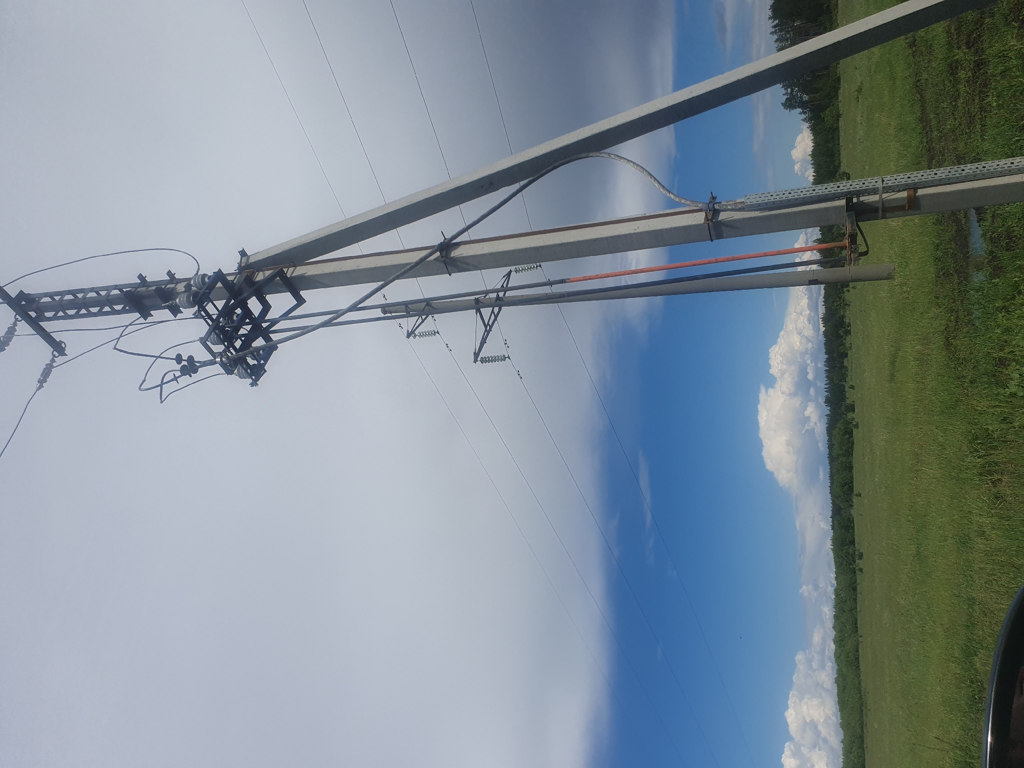 The image shows a utility pole in Архангельское, Russia. The sky is visible with a clear blue gradient and cumulonimbus clouds over a green field. The utility pole is in the left half of the picture and has wires hanging from insulators.