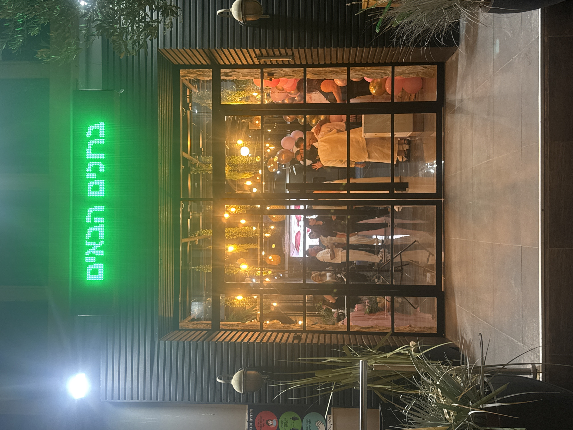 This is a medium shot of the entrance to a building with a green LED sign above the door. The sign displays Hebrew text. The entrance features large glass doors and windows, framed by dark-colored corrugated metal siding on the sides and above the glass. Inside, the space is illuminated by warm-toned string lights and decorative hanging plants, suggesting an indoor event. Several people are visible through the glass, some in formal attire, and there are balloons present, indicating a celebration. In the foreground, to the left, is a large potted plant with tall, thin green leaves. To the far left of the image, a sign with circles and text in Hebrew is attached to the wall. The ground outside the entrance is paved with large, dark tiles. The overall impression is of an evening event at a business or venue in Migdal HaEmek, Israel.