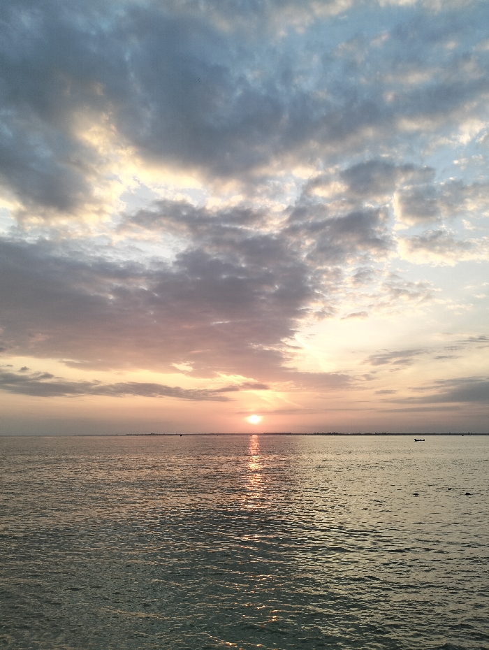 A tranquil sunset scene features a calm ocean with gentle ripples reflecting the sun's radiant path. Above, a sky filled with a mix of white, gray, and blue clouds is illuminated by the setting sun, creating a soft, warm glow. The horizon line is barely visible, marked by faint landmasses and a tiny boat, adding depth to the vast expanse of water and sky.
