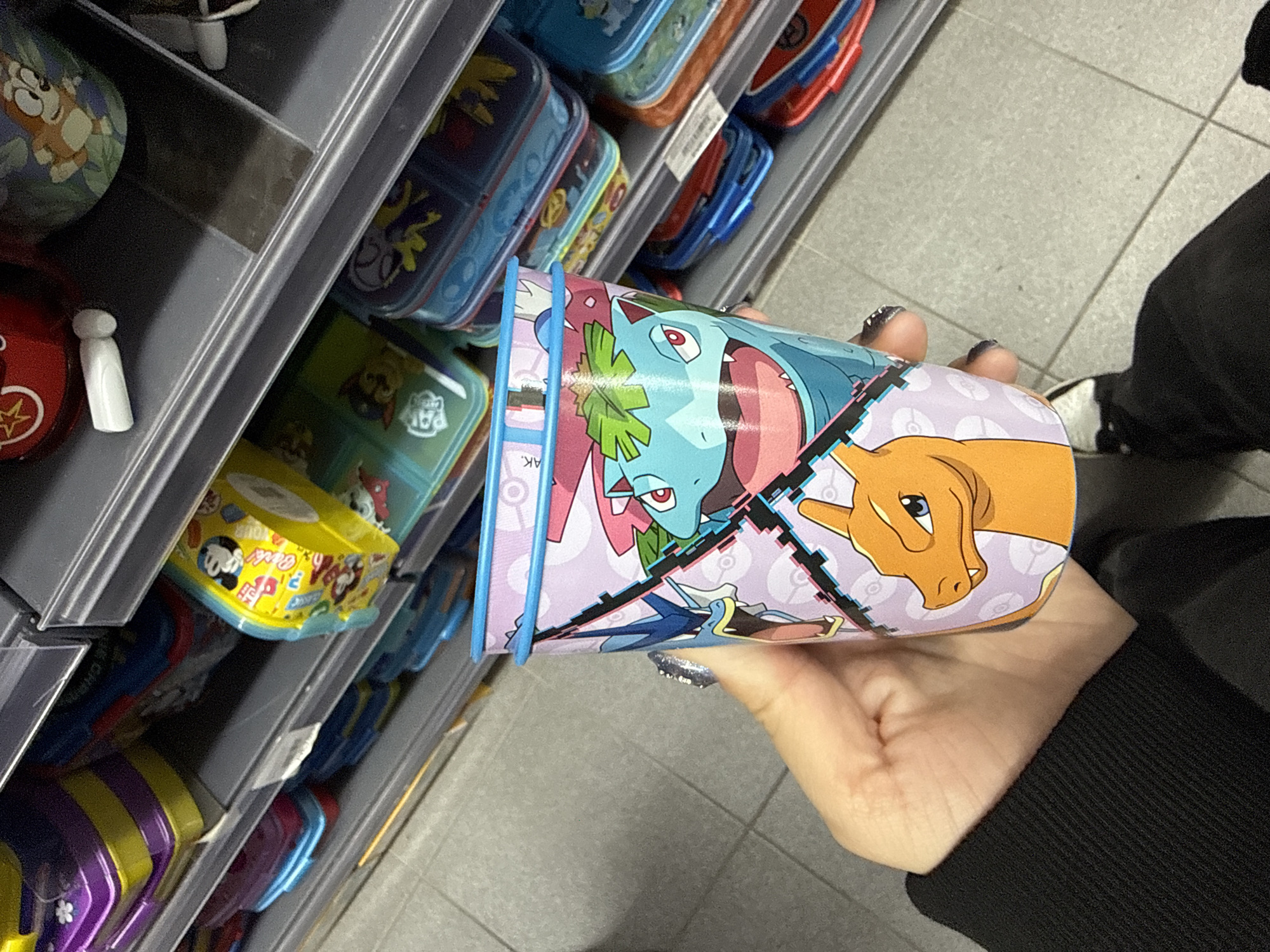 A person stands in a store aisle, holding a cylindrical container with Pokemon characters (Venusaur, Charizard, Blastoise). The aisle is stocked with various lunchboxes. The person is wearing a black long-sleeved shirt and black pants, with white and black shoes partially visible. The location is likely a retail store and not specifically identified as Madrid, Spain.