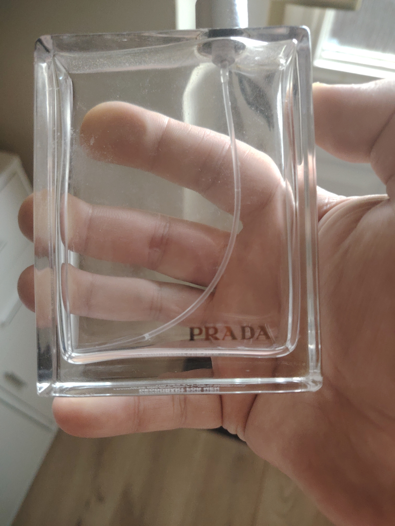 A hand is holding a clear glass rectangular bottle of Prada L'Homme fragrance. The bottle is partially transparent, allowing the fingers and thumb holding it to be visible through the glass. The Prada logo is embossed in the center of the bottle. The background appears to be an indoor setting with a window and furniture visible. The lighting suggests it is daytime. The image was taken in Huntingdonshire, United Kingdom.