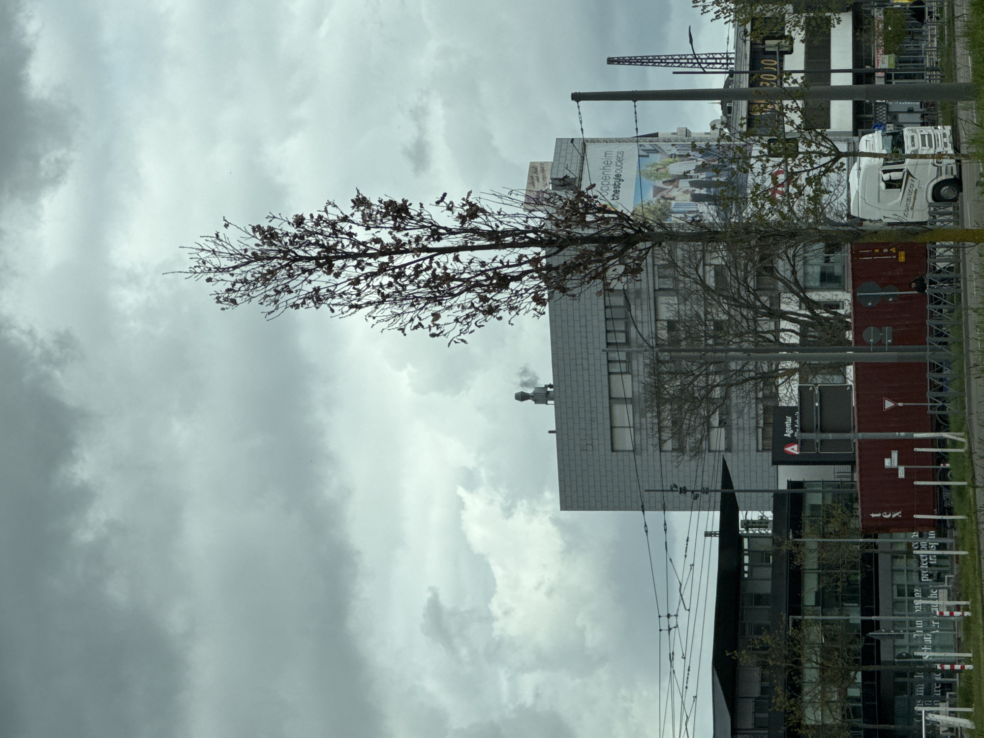 The image depicts a cloudy sky over a streetscape in Kehl, Germany. A tree with bare branches is in the foreground. Behind the tree, there is a multi-story building with windows. Adjacent to it, there's a sign bearing the word "Agentur." The background shows a truck and advertising billboards, and what appear to be construction cranes. Overhead power lines are also visible. The overall scene suggests an urban or industrial area.