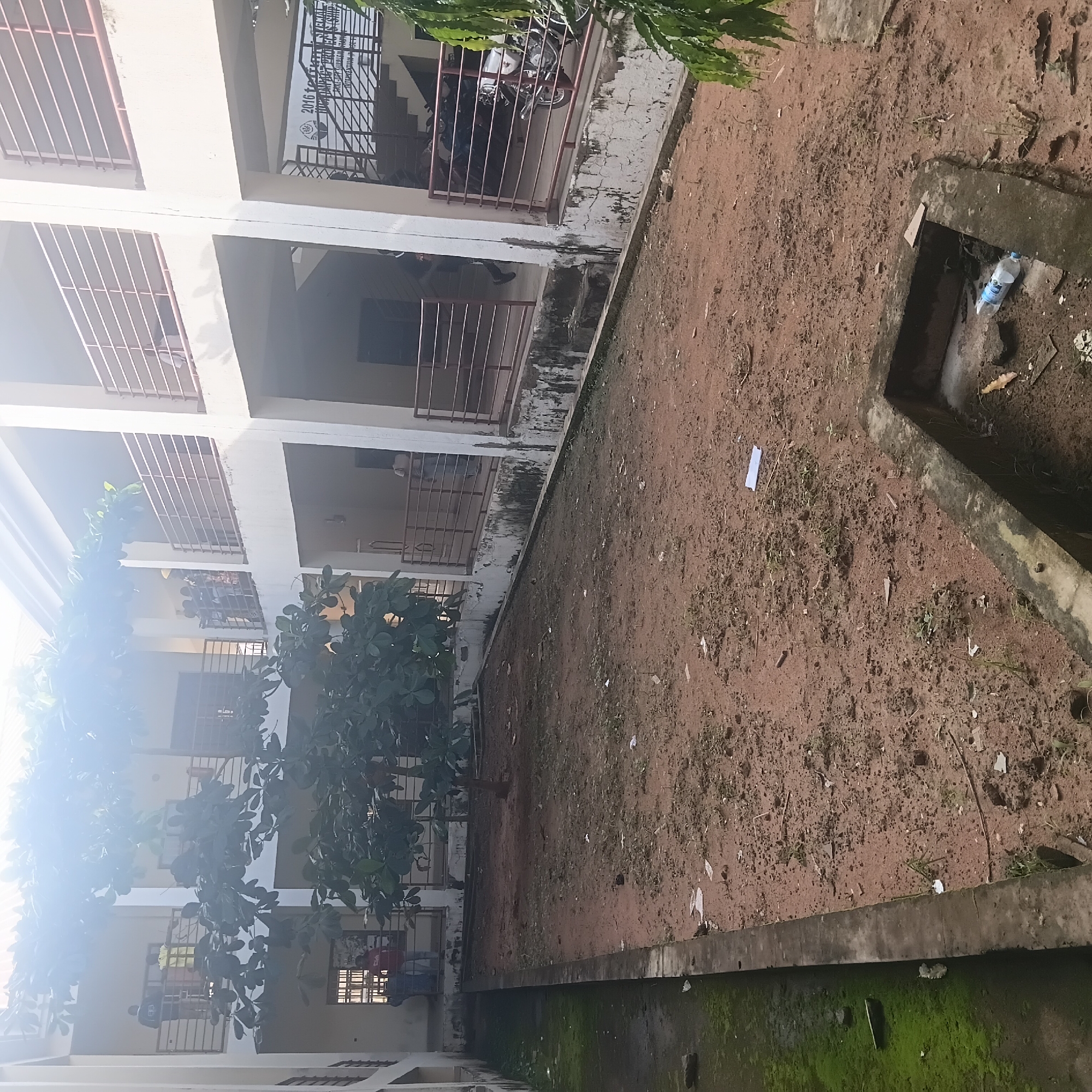 The image depicts an outdoor courtyard area within a multi-story building, likely an educational institution, in Wukari, Nigeria. The courtyard itself is unpaved, with reddish-brown soil and scattered debris, including small pieces of paper and twigs. A concrete-lined drain or channel runs along the left side of the courtyard, partially covered in green moss.
The building has at least two visible levels with balconies or walkways, each lined with metal railings. Several people are visible on the upper levels, appearing to observe the courtyard or interact with each other. On the lower level, near an entrance, a few individuals are standing. In the foreground, a low, concrete structure, possibly a planter or an open drainage basin, contains a discarded plastic water bottle.
A small, leafy tree is centrally located in the courtyard. The building's architecture features white walls and support pillars. In the background, a banner with text, including "2016" and other inscriptions, is visible on a wall near a staircase. Several motorcycles are parked in a covered area on the lower level to the right. The overall impression is of a somewhat neglected but functional urban setting, characteristic of a school or public building in Wukari.