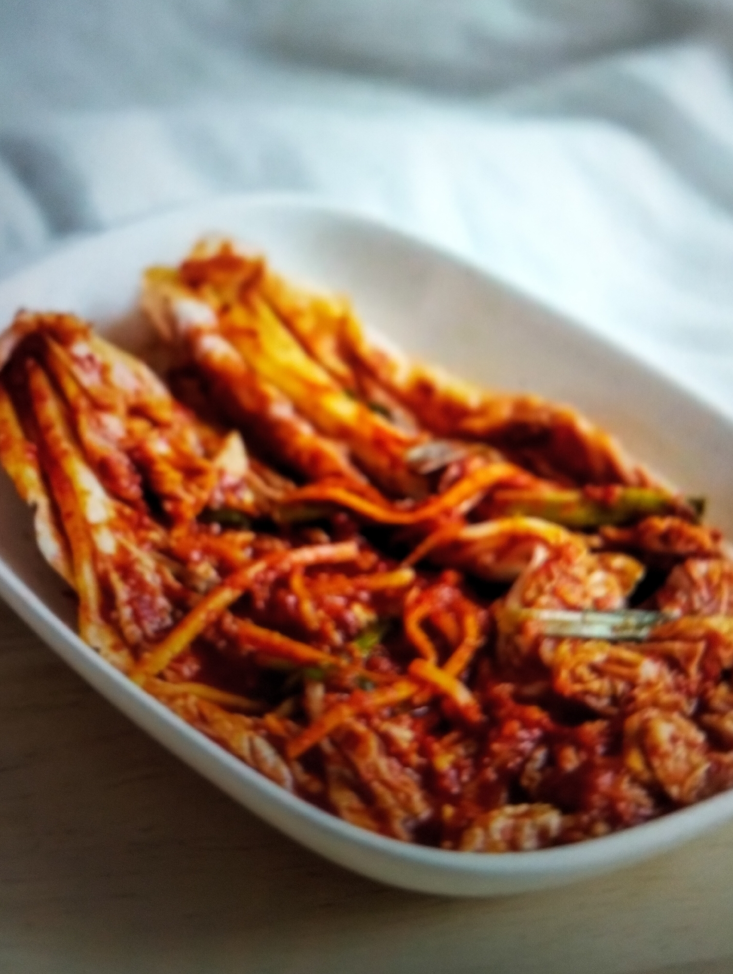 A white ceramic dish filled with kimchi is presented on a wooden surface. The kimchi, a fermented Korean dish, is characterized by its vibrant red hue, a result of gochugaru (Korean chili flakes) and other seasonings. The dish appears to be partially filled, with the kimchi arranged in layers, its leaves showing a slightly wilted texture. The background consists of a softly draped white cloth, creating a gentle, blurred contrast to the textured kimchi. The lighting appears diffused, casting a soft glow on the subject. There is no indication of the specific location in Payagaboh, Indonesia, or any discernible actions, interactions, or individuals in the image.