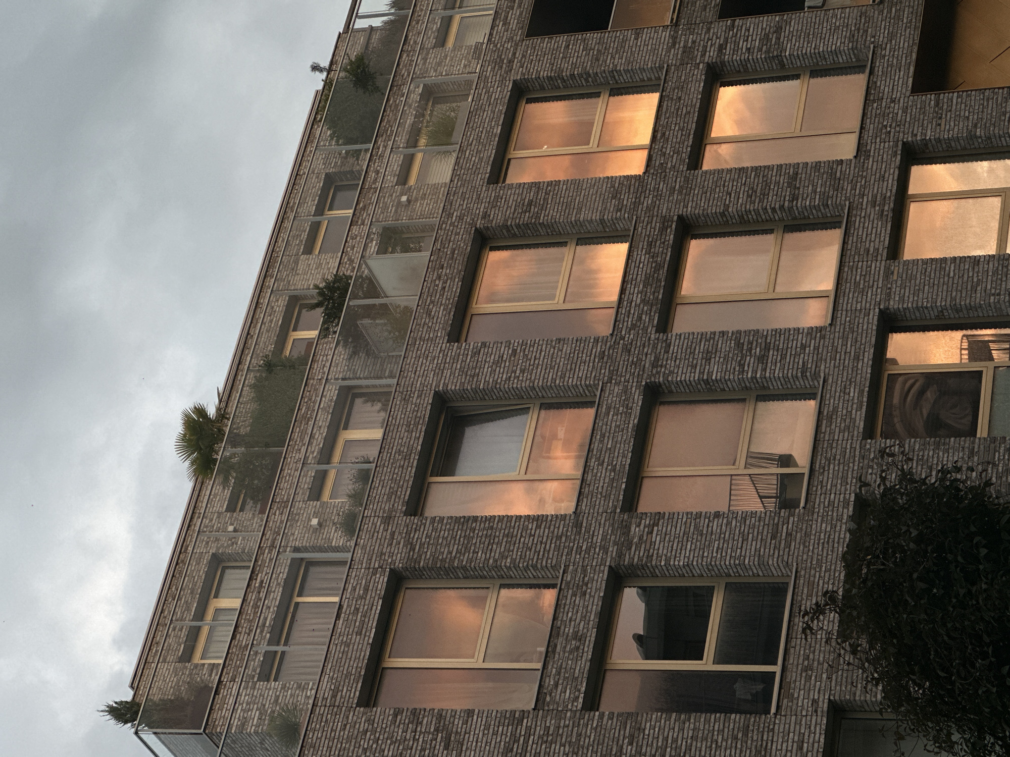 A multi-story apartment building with a brick facade and large windows is pictured in Amsterdam, Netherlands. The windows reflect the orange and pink hues of a sunset sky, creating a warm glow on the building's exterior. Balconies with glass railings and lush greenery are visible on the upper floors, suggesting a modern urban living space. The sky is overcast with dramatic clouds, adding to the atmospheric quality of the scene. A dark, dense bush is positioned in the lower left corner of the image, partially obscuring the building's foundation.