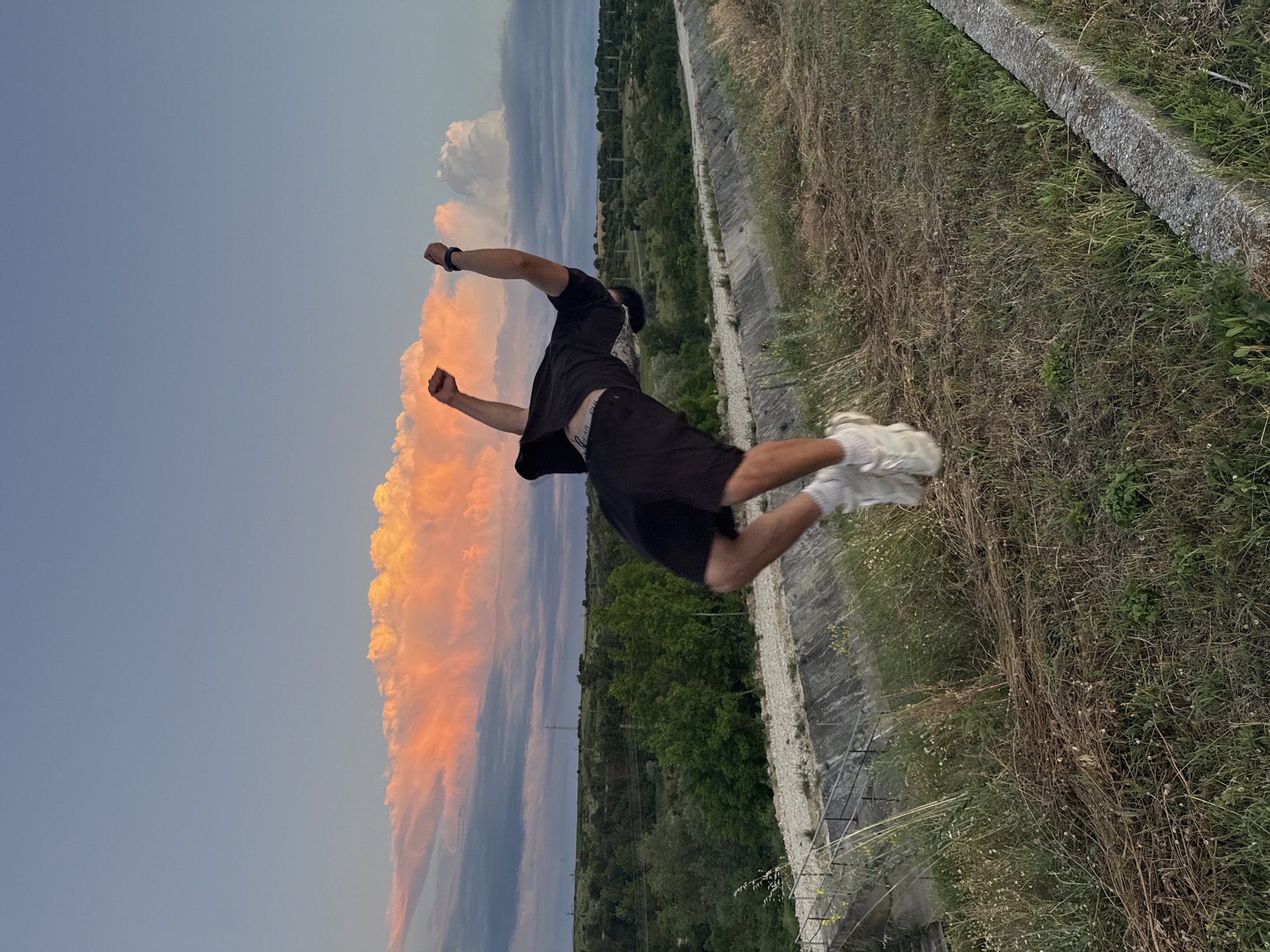 In the city of Shumen, Bulgaria, at dusk, a young man performs a backflip on a grassy embankment. He is wearing a black t-shirt, black shorts, and white athletic shoes. His body is arched backward, arms extended upwards, mid-air. The sky is a blend of pale blue and dramatic sunset colors, with large, illuminated clouds and darker storm clouds. A concrete structure, possibly part of an irrigation channel or road, runs along the middle ground, bordered by dry grass and scattered greenery. Beyond this, a line of trees and vegetation stretches across the horizon, with utility poles visible in the distance. The scene is illuminated by the warm, fading light of the setting sun.