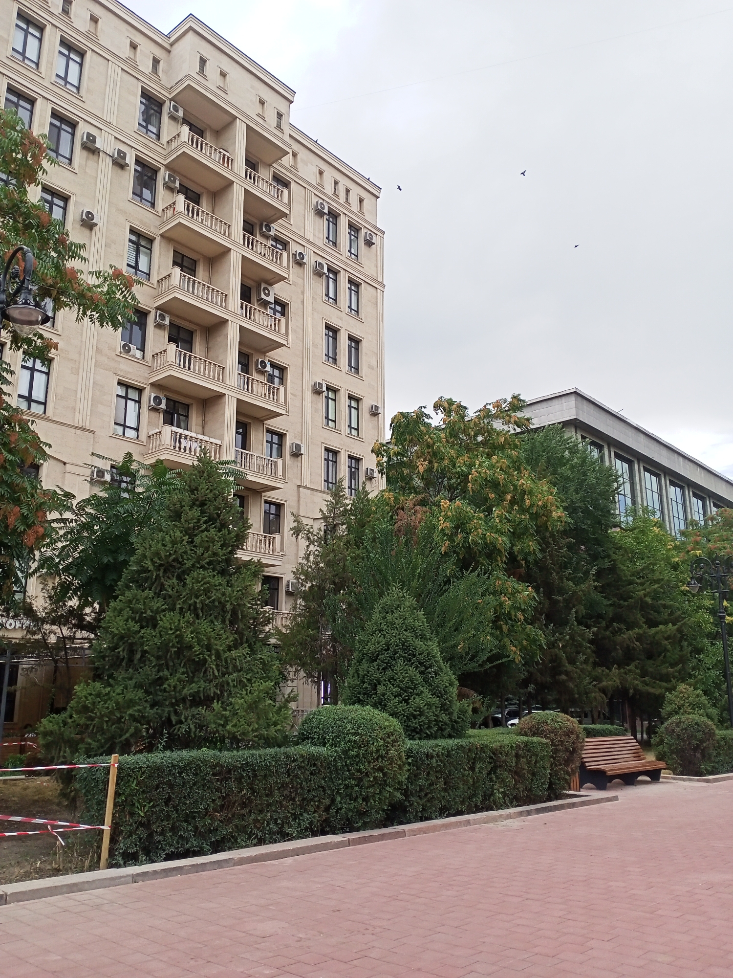 The image shows a building with multiple floors, characterized by its beige facade and numerous balconies with ornamental railings. Several air conditioning units are visible mounted on the exterior walls of the building. In front of the building, there is a pathway paved with red bricks, bordered by a low wall and manicured hedges. A park bench made of wood and dark metal is situated amidst greenery, including evergreen trees and bushes. The sky is overcast with a few birds flying in the distance. The scene appears to be located in Bishkek City, Kyrgyzstan, likely a residential or commercial area with public green spaces. There are no people visible in the image.