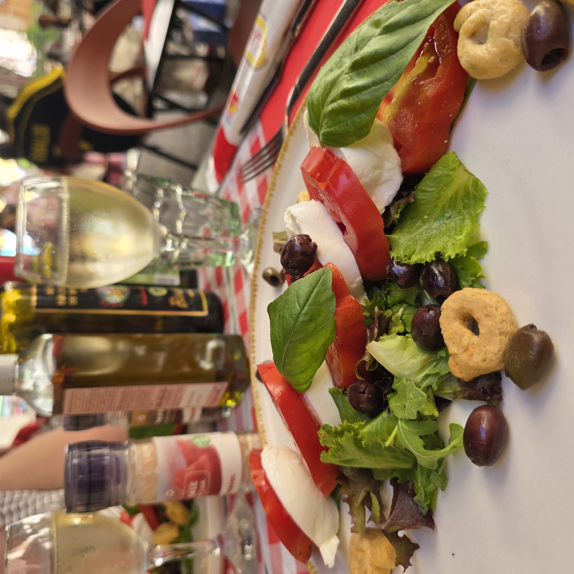 This is a close-up, eye-level shot of a Caprese salad on a white plate, likely at an outdoor cafe in Menton, France. The salad features sliced tomatoes, fresh mozzarella, green lettuce, and Kalamata olives, garnished with fresh basil leaves. Two taralli, a type of Italian cracker, are visible on the plate. In the background, a bottle of olive oil, a shaker of pink salt, and a glass of white wine are present on a red and white checkered tablecloth. The soft focus background suggests other patrons and outdoor seating are nearby.