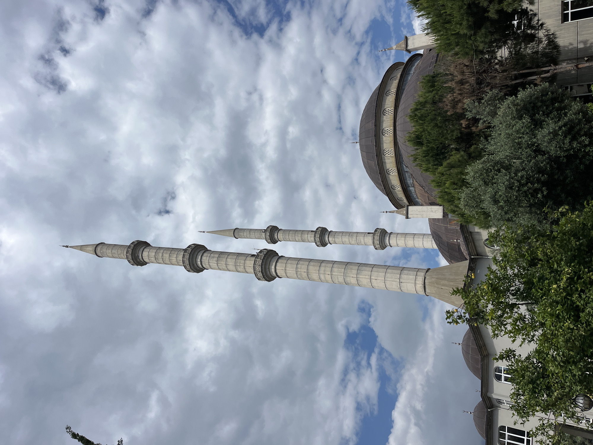 The image shows the exterior of a mosque in Alanya, Turkey. Two tall, slender minarets are prominently visible against a cloudy sky with patches of blue. The minarets are cylindrical and have ornate, ring-like structures around their shafts. The mosque's dome is located to the right, partially obscured by trees. Parts of the building's facade are visible, including windows. The architecture suggests a modern design. There are no people visible in the frame.