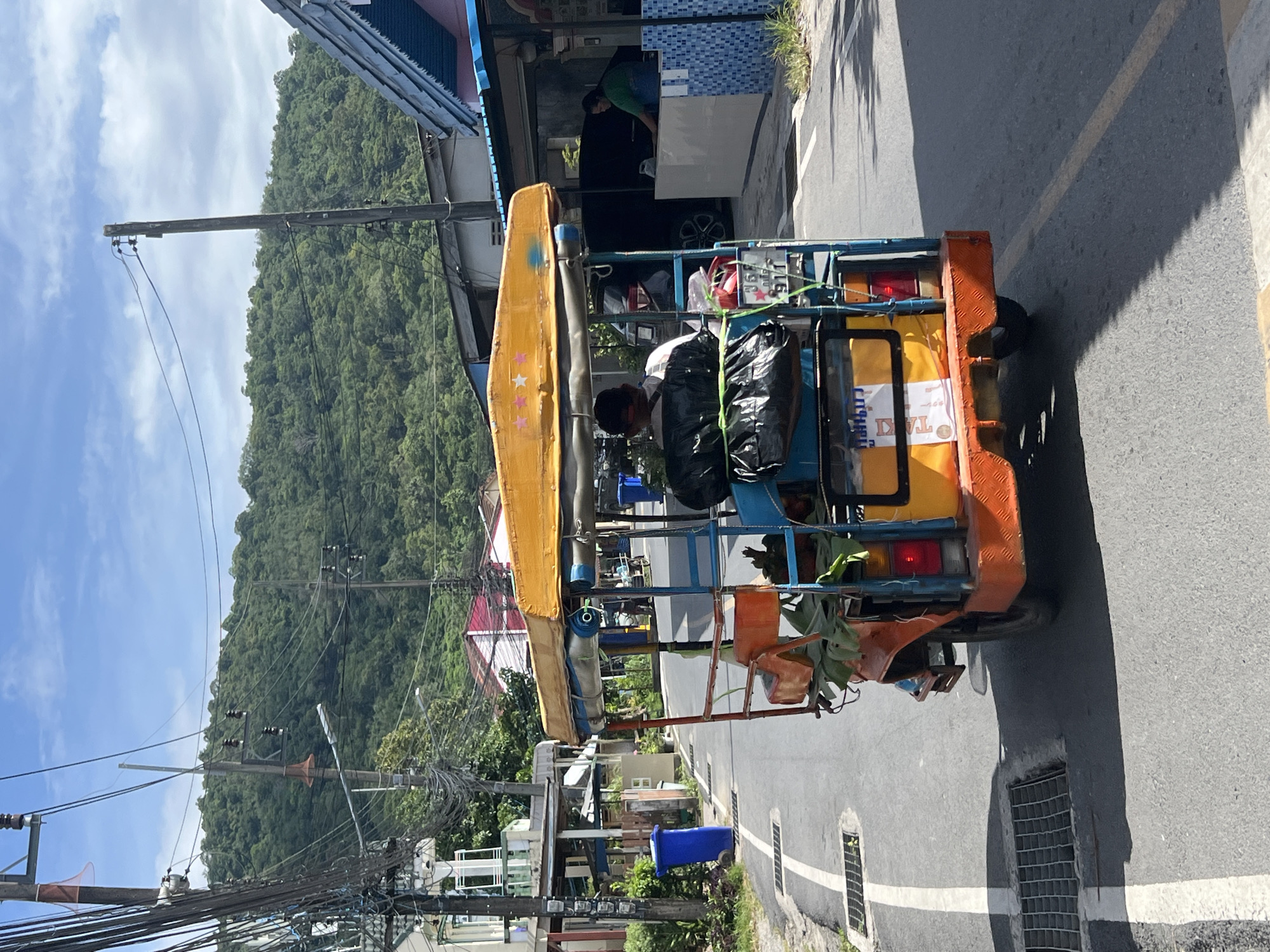 In Choeng Thale, Thailand, a brightly colored tuk-tuk, predominantly orange and yellow with blue accents, is traveling down a paved road lined with buildings and vegetation. The tuk-tuk is carrying two individuals in the back; one appears to be a passenger and the other a driver or operator. The vehicle has a yellow canopy adorned with three pink stars. Bags and foliage are visible in the back of the tuk-tuk. On the right side of the road, a woman in a green shirt is seen interacting with a counter in front of a building. The street is marked with white lines and has a drainage grate on the left. In the background, a lush, green, tree-covered hill rises under a partially cloudy blue sky. Utility poles with numerous wires are also prominent features of the environment.