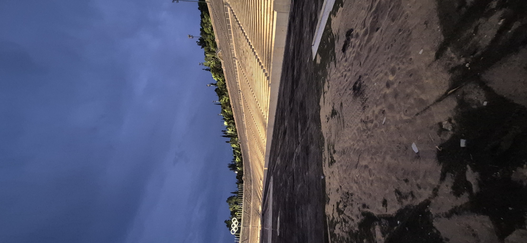 The image shows an exterior, ground-level view of the Panathenaic Stadium in Athens, Greece, at dusk. The stadium's marble seating is illuminated by artificial lights, stretching upwards along the curved sides of the venue. A large Olympic rings symbol is visible on a pole on the left side of the stadium. The foreground is a dirt and grass area, appearing somewhat unkempt, with scattered debris. The sky above is a deep, cloudy blue, indicating the transition from day to night. Trees and lights are visible along the upper rim of the stadium. No people are present in the image.