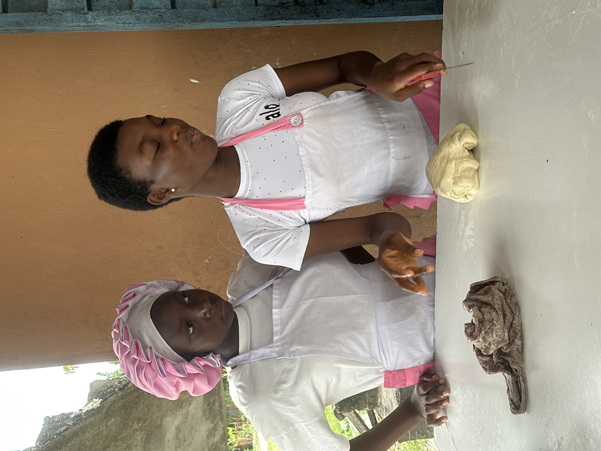 In Badagry, Nigeria, two young women are engaged in a culinary activity, likely preparing dough. The woman on the left, wearing a pink bonnet and a white apron over a white t-shirt, has her hands resting on a white surface. The woman on the right, with short, dark hair, also wears a white apron over a white t-shirt with a pink trim and the word "alo" printed on it. She is actively working with a lump of light-yellow dough, using a red-handled knife to cut into it. In the foreground, a crumpled piece of fabric lies on the white surface. The background features an orange wall and some greenery visible to the left.