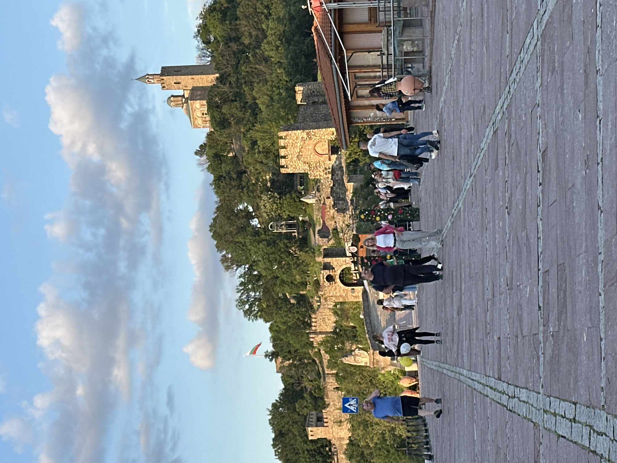 The image depicts a scene in Veliko Tarnovo, Bulgaria. A paved pedestrian area extends into the foreground, with numerous people walking and standing. In the background, a fortress is situated atop a hill, partly obscured by dense trees. The fortress features stone walls and a church or tower. A flag is visible, partially obscured by the foliage. The sky is partly cloudy. The scene is captured in daylight with what appears to be late-afternoon or early-evening lighting.