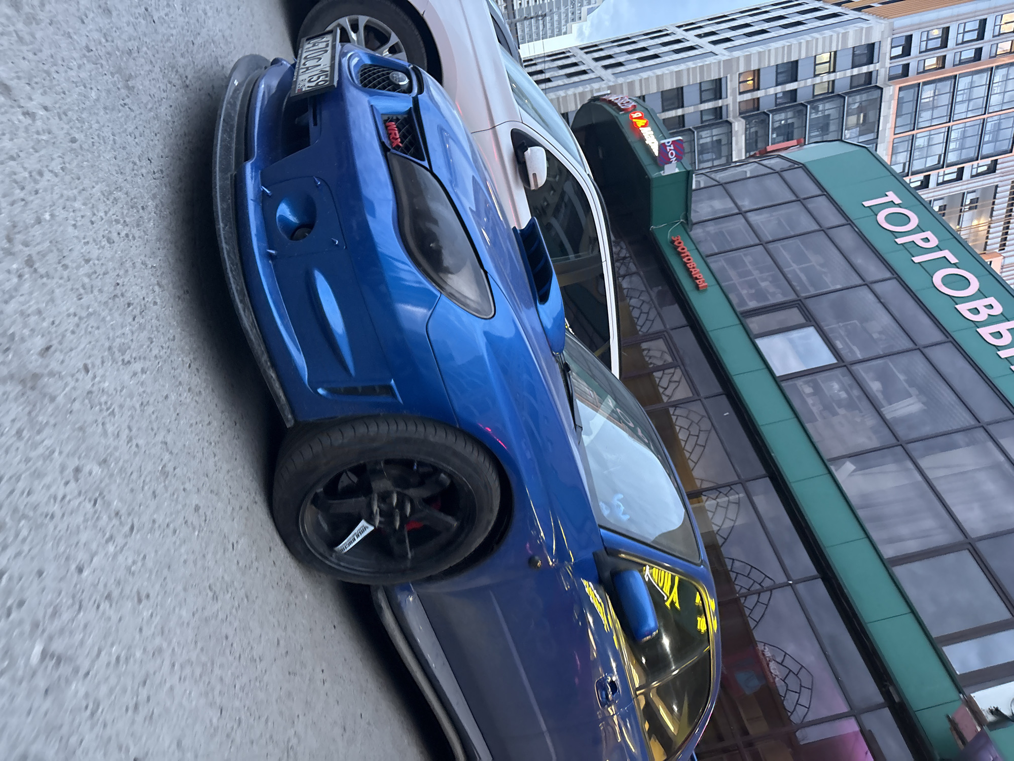 The image shows a blue Subaru WRX parked on a city street in Perm, Russia. The car has blacked-out headlights and a front splitter. A white vehicle is partially visible behind the Subaru. The background features a multi-story building with a green awning displaying the word "ТОРГОВЫЙ" (shopping) in pink Cyrillic letters. Another sign with "ПРОДТОВАРЫ" (food products) is visible on the awning. The building's facade has a modern design with large windows. The lighting indicates it is likely daytime or dusk.