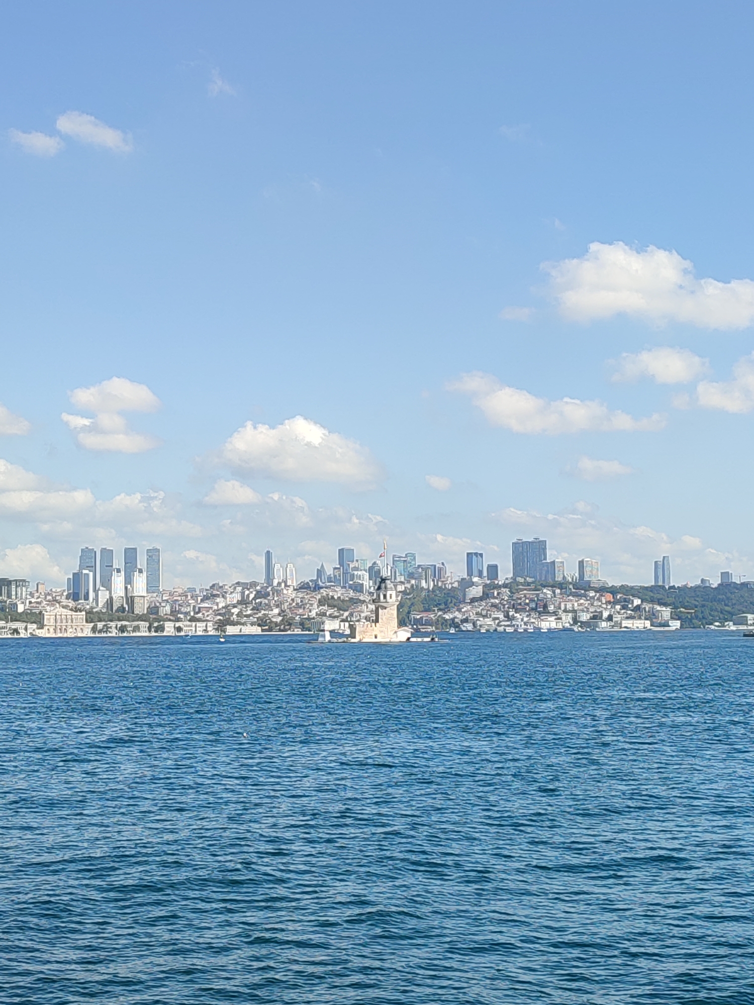 This is a wide-angle, medium-shot photograph taken from the water, looking towards the Üsküdar district of Istanbul, Turkey. The scene is dominated by the Bosphorus strait in the foreground, its surface a vibrant blue with subtle ripples and wave patterns. In the middle ground, the Üsküdar skyline stretches across the horizon, a mix of historic and modern architecture.
Prominently featured is the Maiden's Tower (Kız Kulesi), a historic structure situated on a small islet just off the coast of Üsküdar. The tower is a focal point, its stone construction and distinctive cylindrical shape clearly visible. Behind it, a dense urban landscape unfolds, with buildings of varying heights and architectural styles. Several modern skyscrapers, characterized by their glass facades and geometric shapes, stand out against the older, lower-rise structures. The buildings are clustered together, creating a dynamic silhouette against the sky.
The background is a clear blue sky with scattered white clouds, adding depth and a sense of a bright, sunny day. The lighting suggests midday or early afternoon. The overall impression is a picturesque view of Istanbul's waterfront, showcasing the harmonious blend of its historical heritage and contemporary development. There are no visible people in the frame, and the water appears calm, with a few small boats visible in the distance.