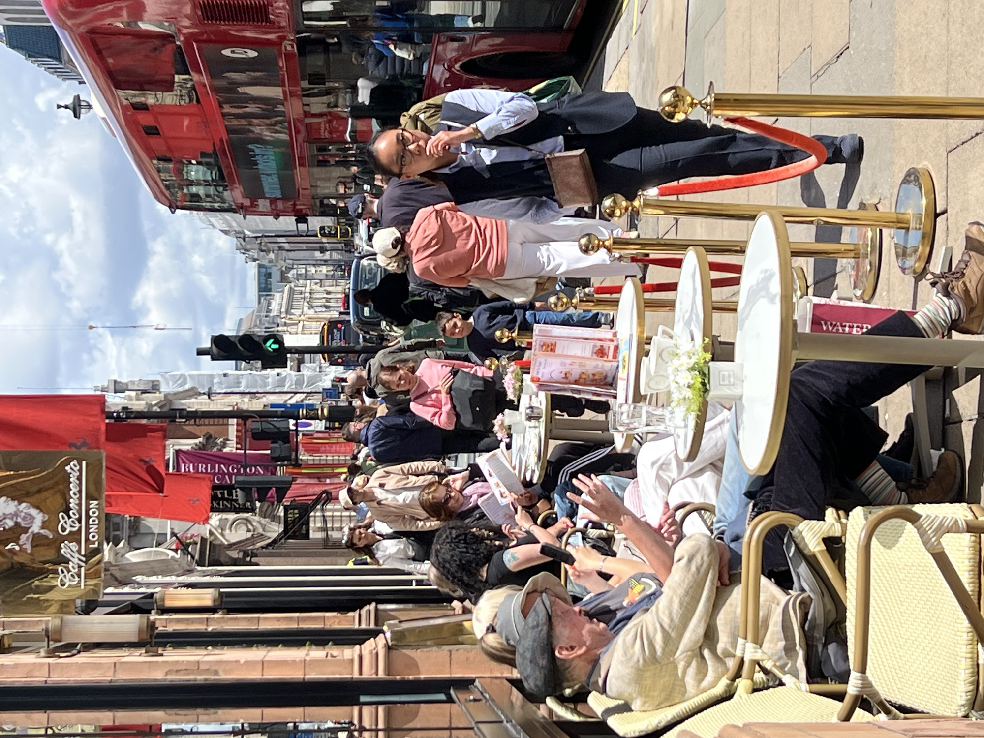 The image depicts a street scene in London, United Kingdom, likely Regent Street, with several people seated at outdoor tables of a cafe. The cafe, identified by signage as "Caffe Concerto London," is situated on the left side of the frame, featuring traditional building architecture with large windows and decorative elements. In the background, a red double-decker bus is visible, a common sight in London.
In the foreground, individuals are engaged in various activities. A man in a light-colored jacket and cap is seated at a table, looking at his phone. Adjacent to him, other patrons are reading, talking, and enjoying their time outdoors. The tables are set with white tablecloths, cups, and what appear to be pastries.
The environment is characterized by a busy urban setting with pedestrian traffic and vehicles. The sidewalk is paved with stone tiles, and decorative stanchions with red ropes delineate the outdoor seating area of the cafe. The day appears to be sunny with some clouds in the sky, suggesting pleasant weather. The overall atmosphere conveys a sense of casual dining and people-watching in a vibrant city location.