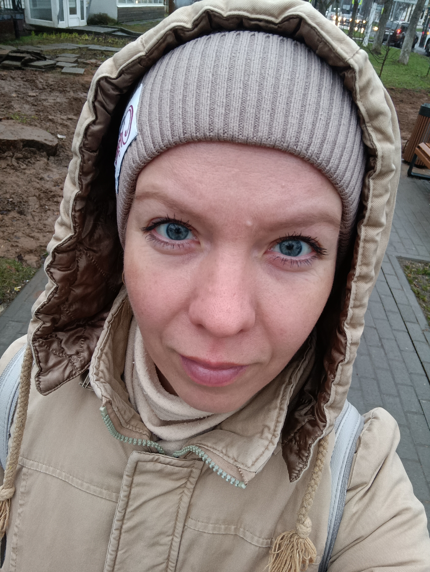 A close-up photograph depicts a woman with fair skin and striking blue eyes. She wears a beige knit hat and a hooded tan jacket, zipped up to the neck. The hood is lined with a brown, quilted material. The woman looks directly at the camera with a slight upward tilt of her head.
The background reveals an urban environment, with a paved walkway visible to the right. A patch of muddy ground with some greenery is seen to the left. Further in the background, blurred trees and buildings can be discerned, suggesting a city park or street scene. The presence of distant vehicles indicates a road nearby. The location is Chaikovsky, Russia.