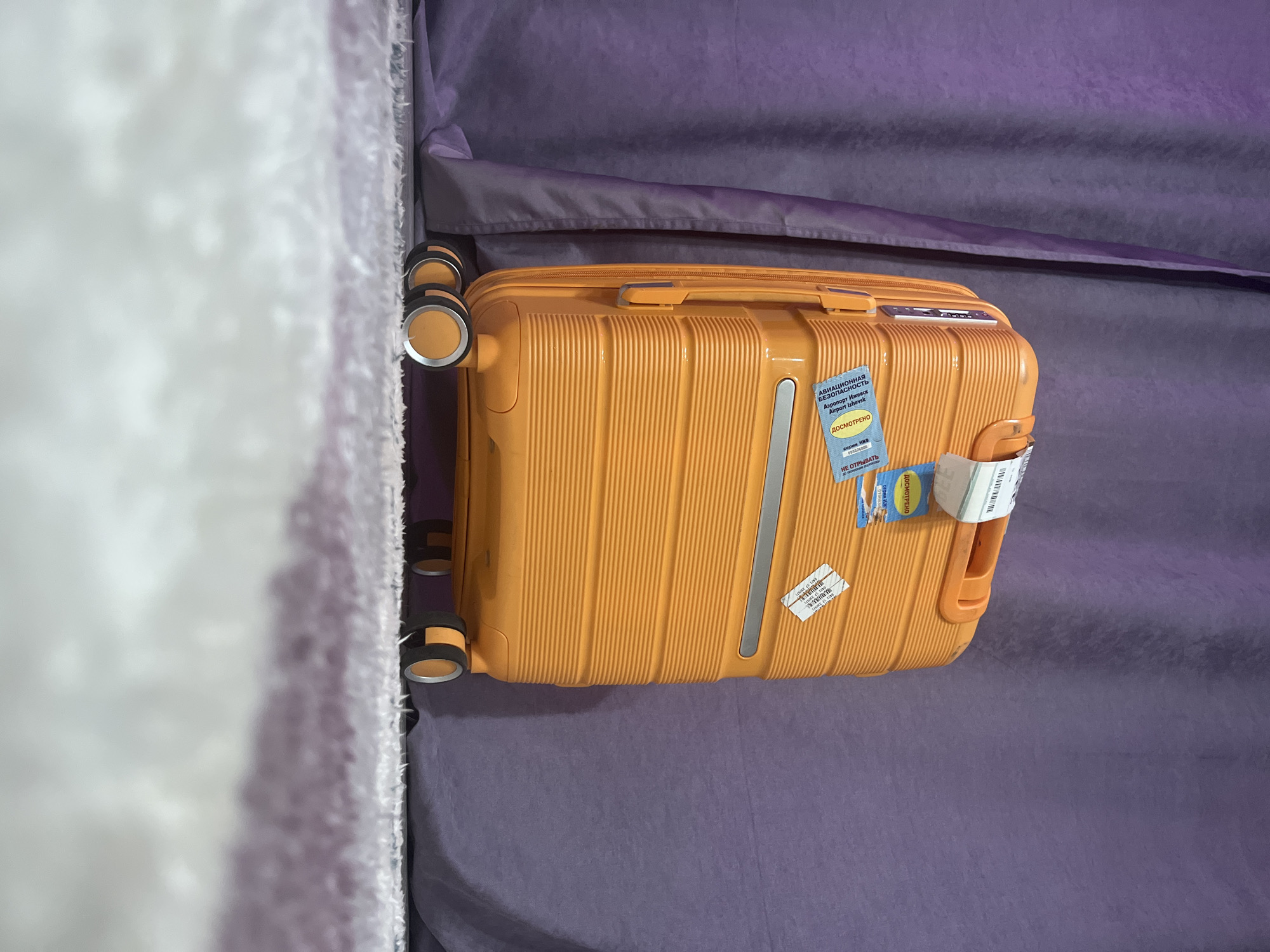 An orange suitcase with airport security stickers rests on a lilac-colored surface, possibly a bed or a flat platform covered with fabric. The suitcase is oriented diagonally in the frame. Part of a white, plush object, possibly a wall or furniture covering, is visible on the left side of the image. The image appears to have been taken in a close-up or medium shot. There is no evidence to confirm the location is Chaikovsky, Russia.