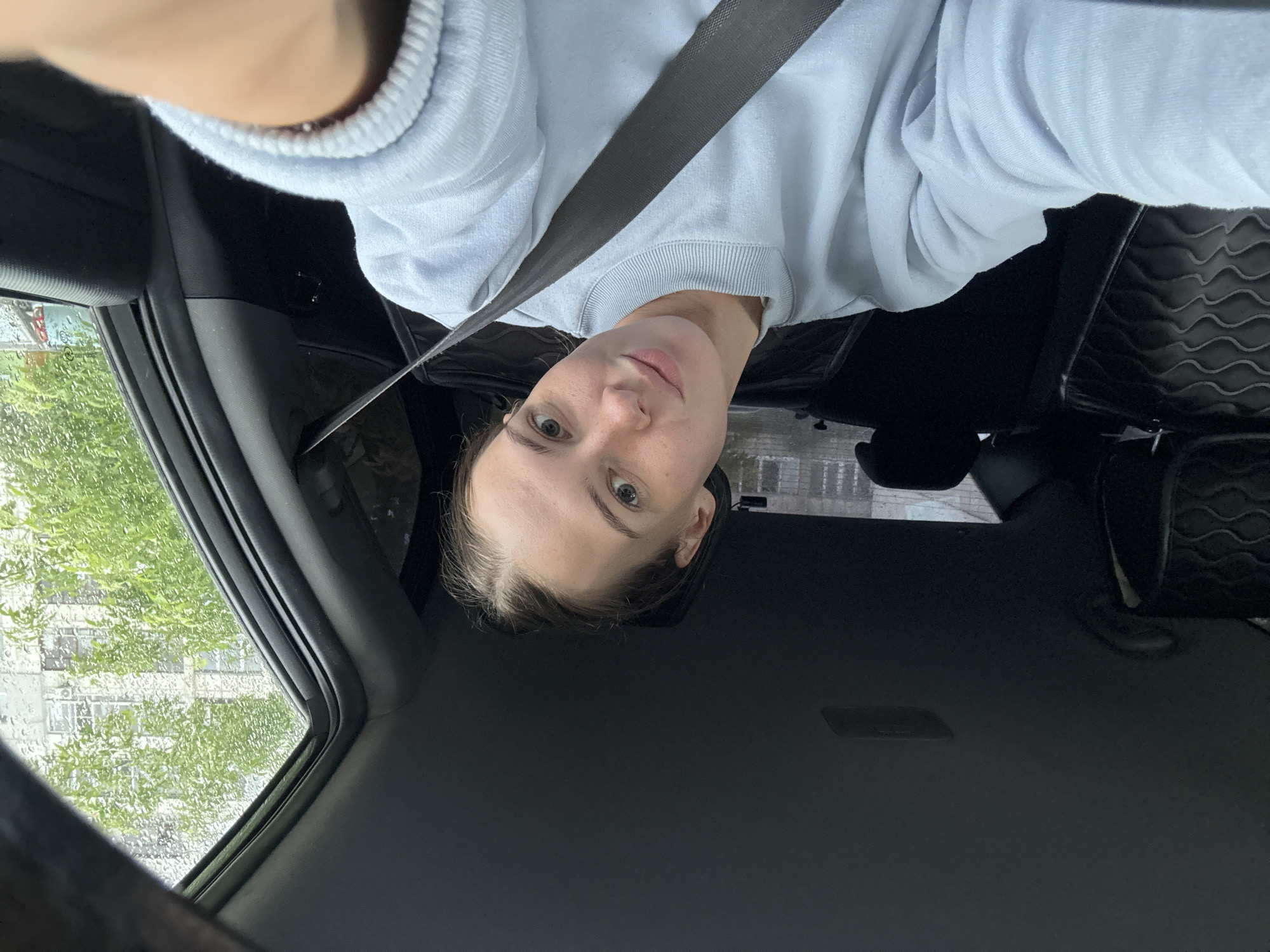 A young woman is taking a selfie inside a car, presumably in the city of Perm, Russia. She is wearing a light blue sweatshirt and a grey seatbelt. The background shows the interior of the car, including the headrests of the seats and the car's ceiling. Through the window, there are trees and a building, suggesting an urban or suburban environment. The weather appears to be rainy or overcast, as evidenced by the water droplets on the window. The woman is looking directly at the camera with a gentle smile.