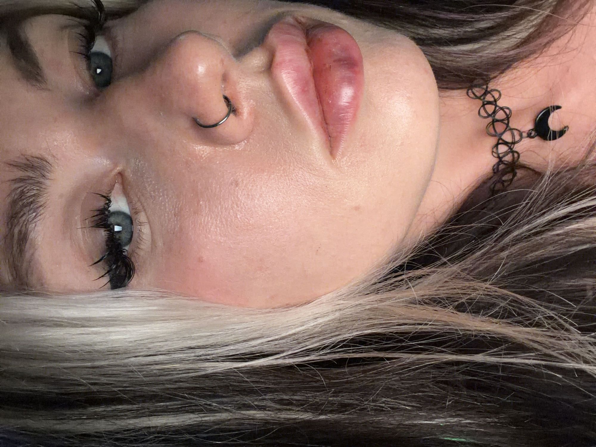 A close-up of a woman's face, angled and slightly tilted. She has fair skin, light blue eyes with dark mascara, and a silver nose ring. Her lips appear swollen and bruised. She wears a black choker necklace with a crescent moon charm. Her hair is dark with streaks of blonde. The background is out of focus. There is no indication of location in Budva, Montenegro.