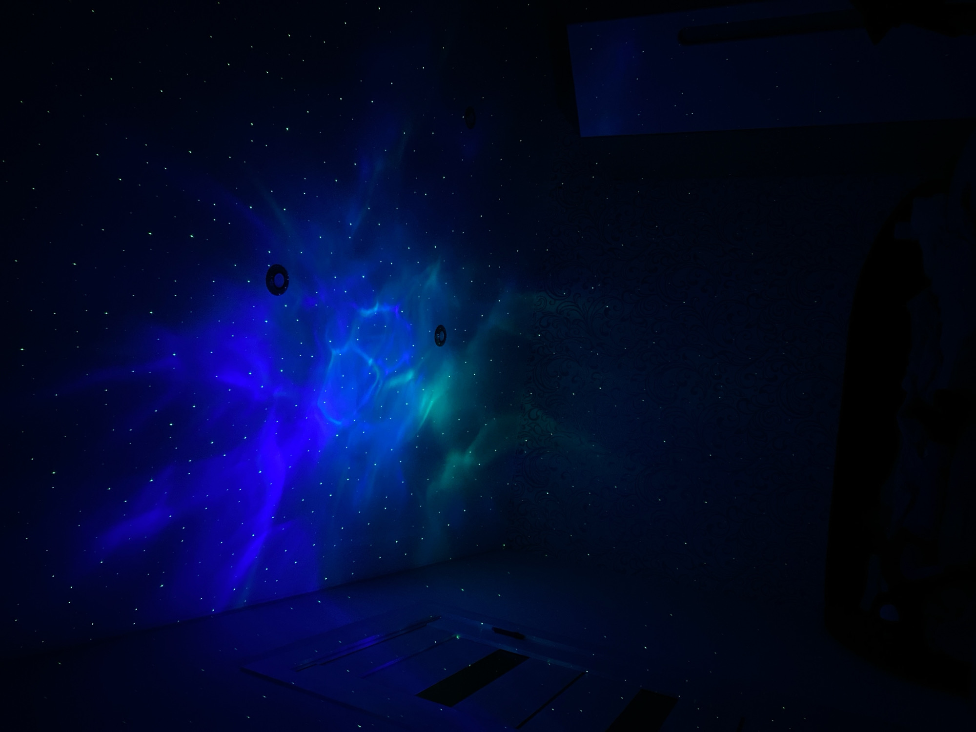 The image depicts a darkened room with a projection of a nebula-like pattern on the walls and ceiling. The projection is primarily blue and green, with small, bright dots resembling stars scattered across the surfaces. There are two circular, dark objects on the wall within the projected nebula. The floor appears to be made of light-colored panels with darker rectangular sections. To the right, a dark figure is vaguely visible. The room is in Markovskii, Russia.