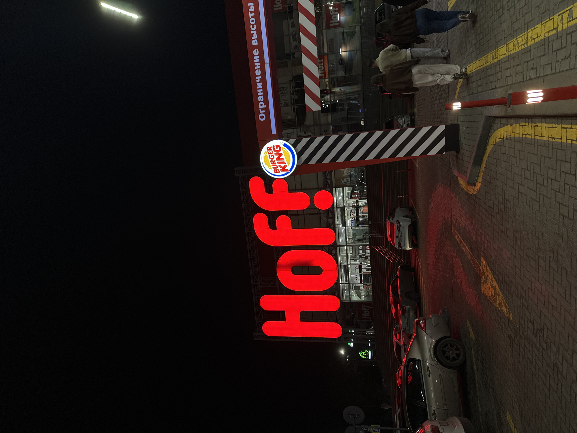 The image shows a nighttime scene at a Burger King and Hoff location in Rostov-on-Don, Russia. The Hoff sign is large and red, positioned prominently next to the Burger King logo. The sign "Ограничение высоты" is visible. People are present near the entrance. Several cars are parked in the parking area. The environment appears to be a standard urban commercial area at night, with artificial lighting illuminating the scene.