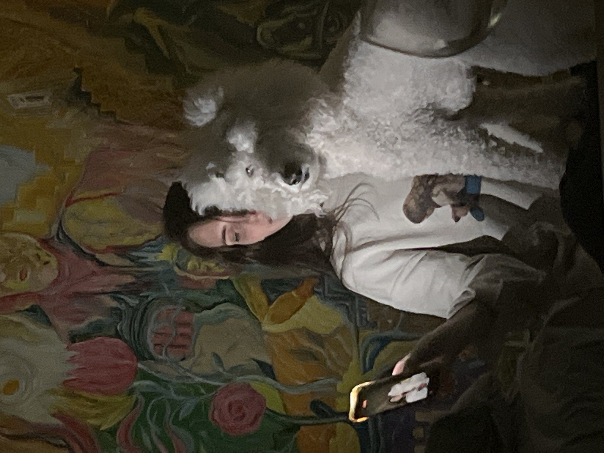 The image depicts a woman lying down next to a white poodle. She is wearing a white t-shirt. Her eyes are closed and the dog is positioned with its head near her face. A person is holding a smartphone in the lower right of the image and appears to be taking a photo. The background is a colorful painting with floral and abstract elements. The location is unknown.