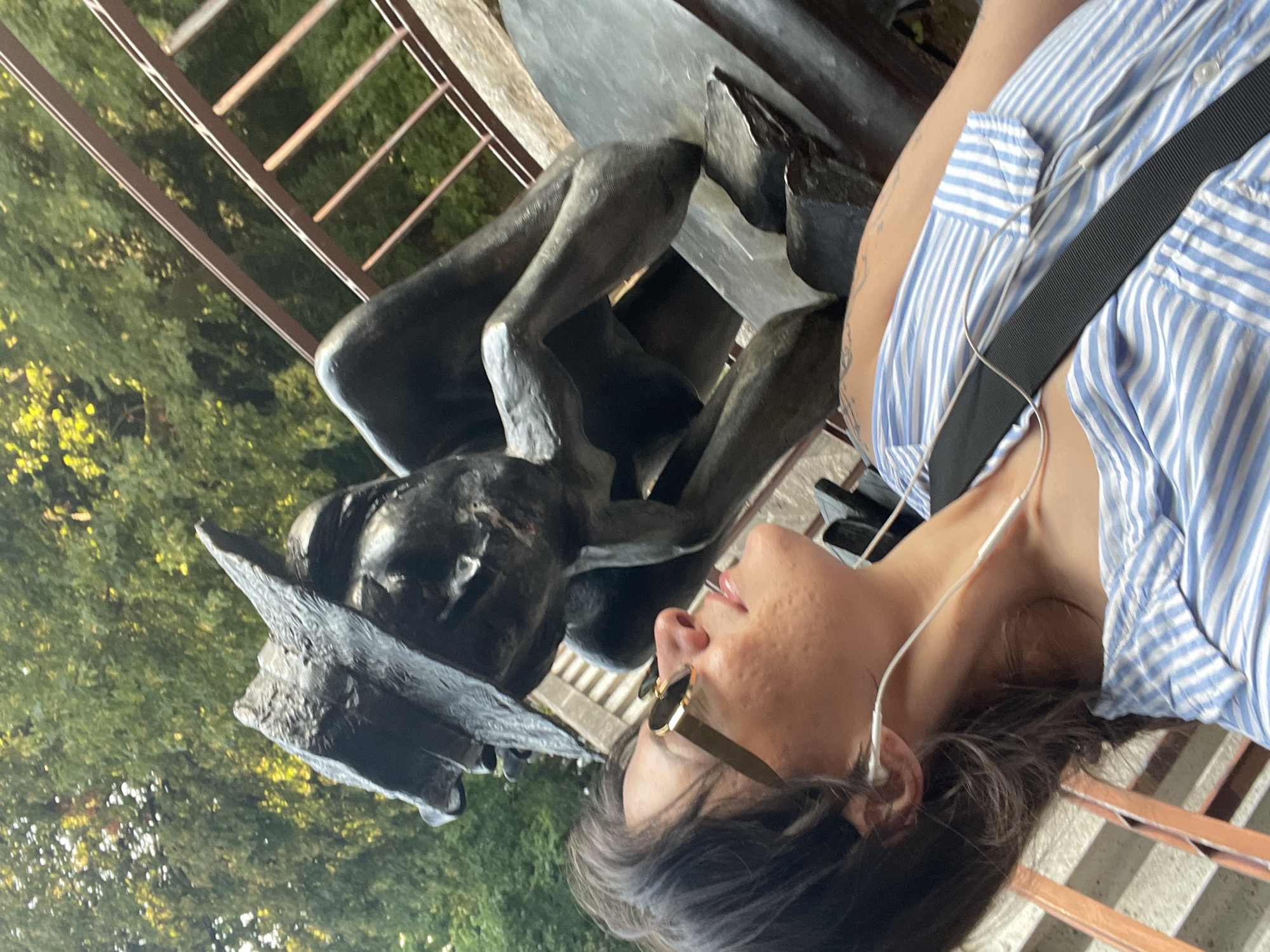 A woman in a blue and white striped shirt and sunglasses looks up at a bronze statue of a contemplative figure. The statue is situated outdoors, with trees and a railing visible in the background. The woman is wearing earbuds. The scene is likely in Iserlohn, Germany.