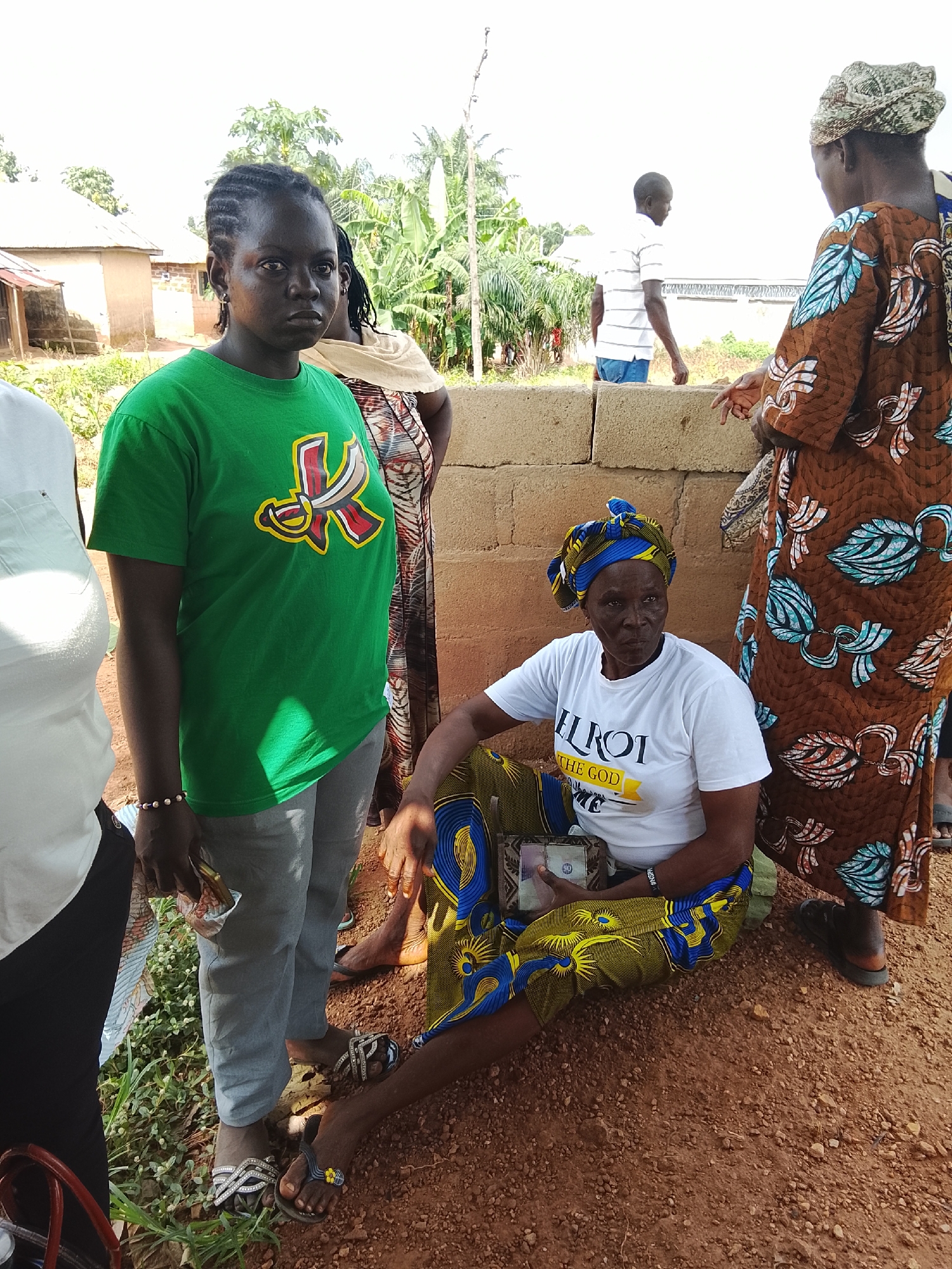 In Akwanga, Nigeria, a group of people stand in an outdoor area with a sandy ground and a brick wall. In the foreground, a young woman wears a green t-shirt with a red and yellow logo and grey pants. Next to her, a woman in a white t-shirt with yellow lettering and a blue and yellow patterned skirt sits on the ground, holding money. To the right, a woman in a brown and blue patterned dress stands. Behind them, a man in a striped white shirt and blue jeans stands near a building. The environment suggests a rural or semi-rural setting, possibly a community gathering.