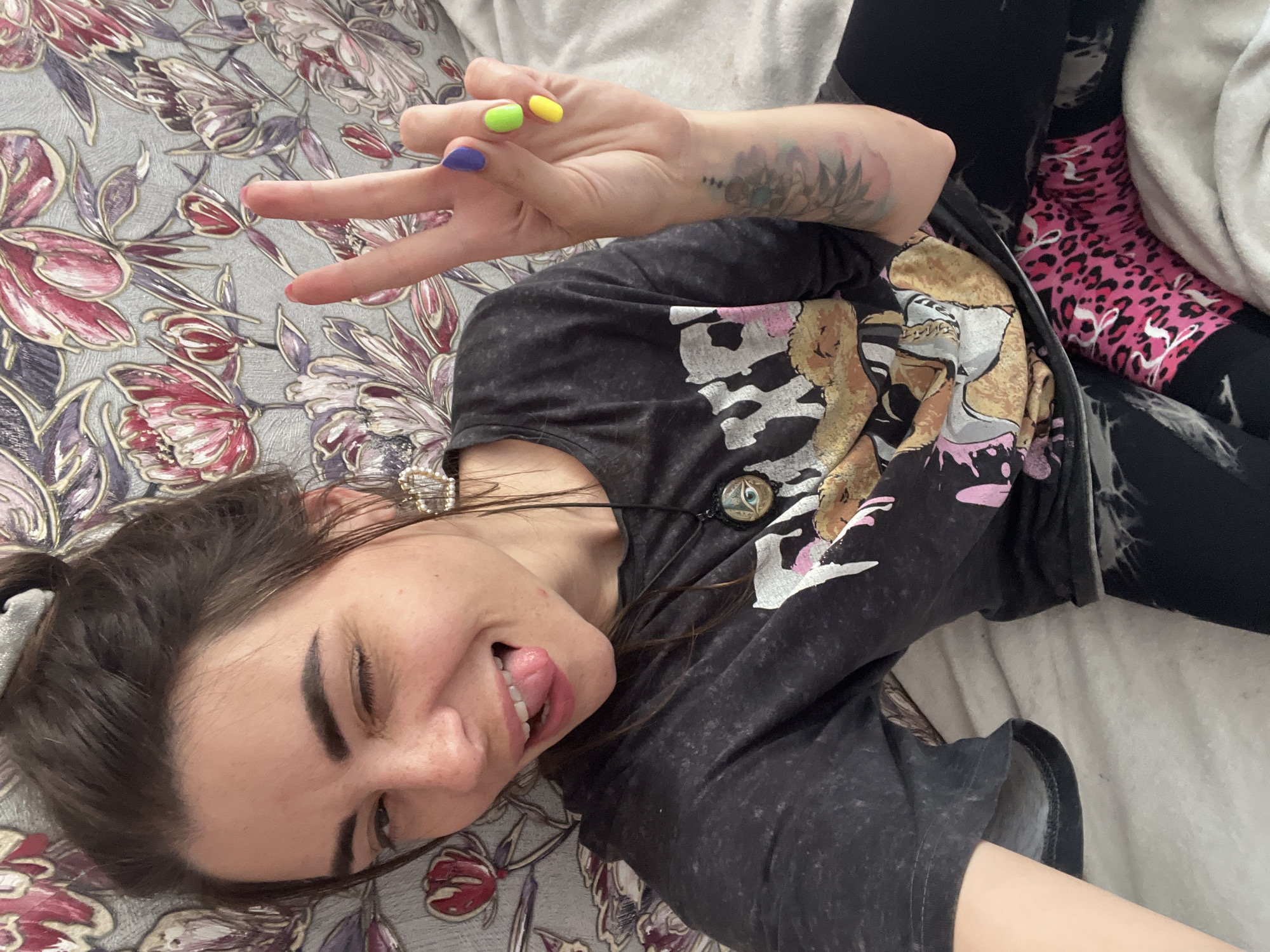 A young woman is lying on a bed, looking at the camera with her tongue sticking out and one eye winked. She is wearing a dark t-shirt with a graphic print and black leggings with bright pink leopard-print socks. She is making a peace sign with her right hand, which has brightly colored fingernails. A patterned tapestry or wallpaper is visible behind her. The image appears to be a selfie taken in a bedroom. There is no information available to determine the location within the city of Kostroma, Russia.