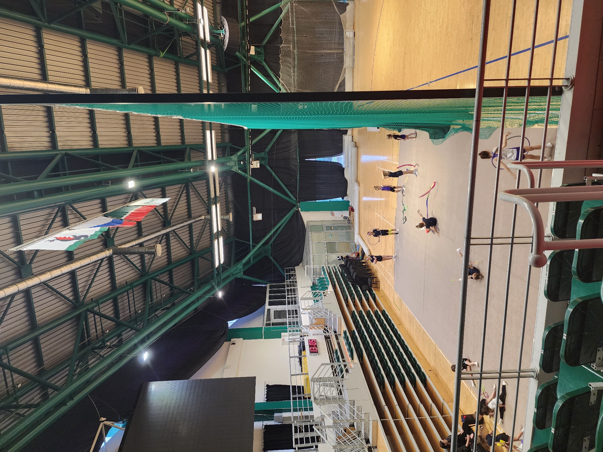 This image captures a gymnasium scene in Kazan, Russia. The space features a light-colored wooden floor, occupied by several individuals engaged in rhythmic gymnastics activities, including routines with ribbons and hoops. Seating arrangements run along the side of the gym. The structure's roof is metal, supported by a network of green beams and trusses. A green net hangs down, and a Russian flag hangs from the ceiling. A digital scoreboard is visible along one wall, near a seating area.